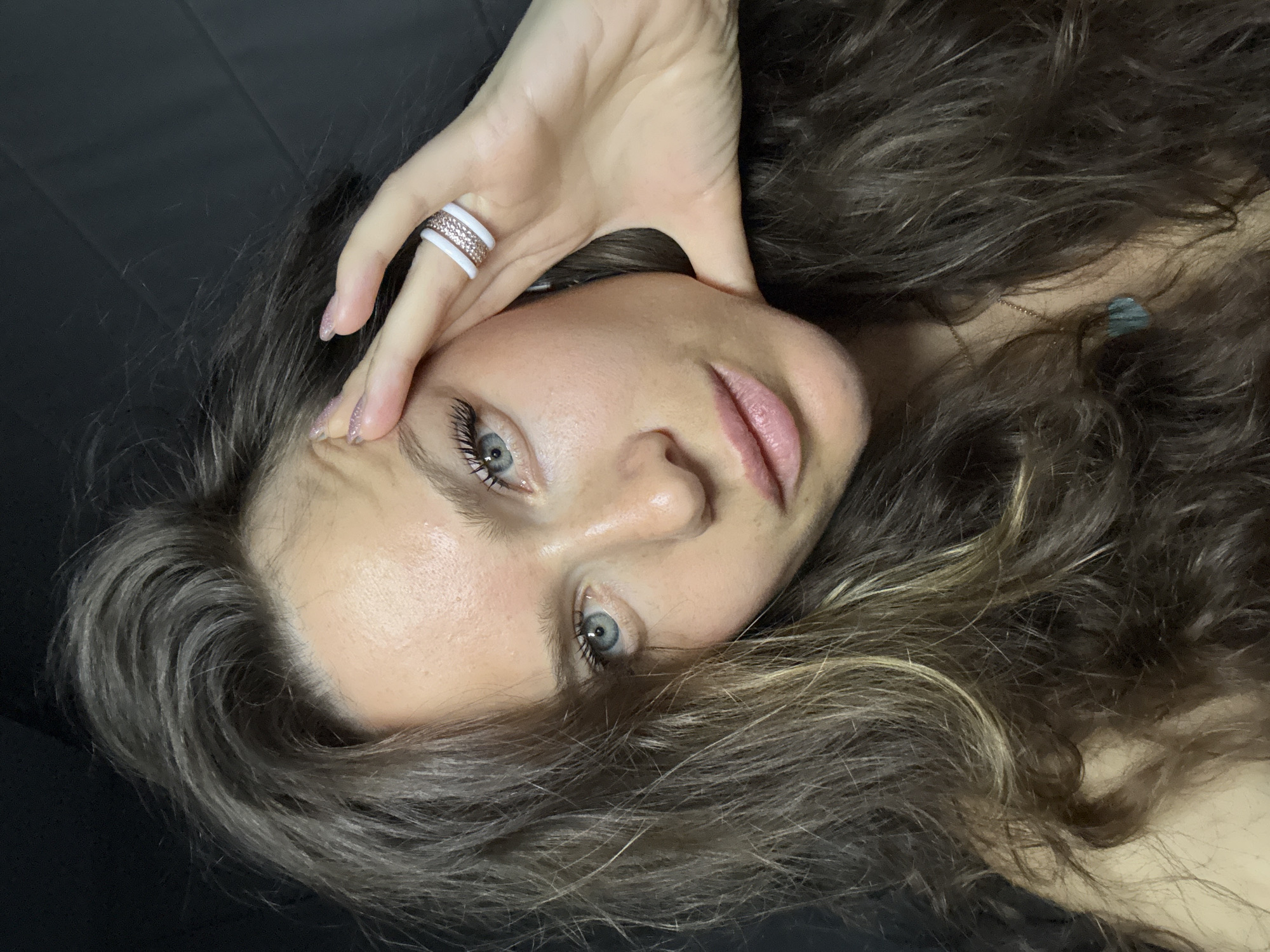 A close-up, angled view of a woman with long, wavy brown hair streaked with blonde highlights. She has light skin, blue eyes, and pink lips. Her right hand is raised, with her fingers touching her forehead near her temple. She is wearing a white ring with a rose gold band containing small stones on her ring finger. A gold chain necklace is partially visible amid her hair. The background is dark and out of focus. The location is not discernible from the image alone.