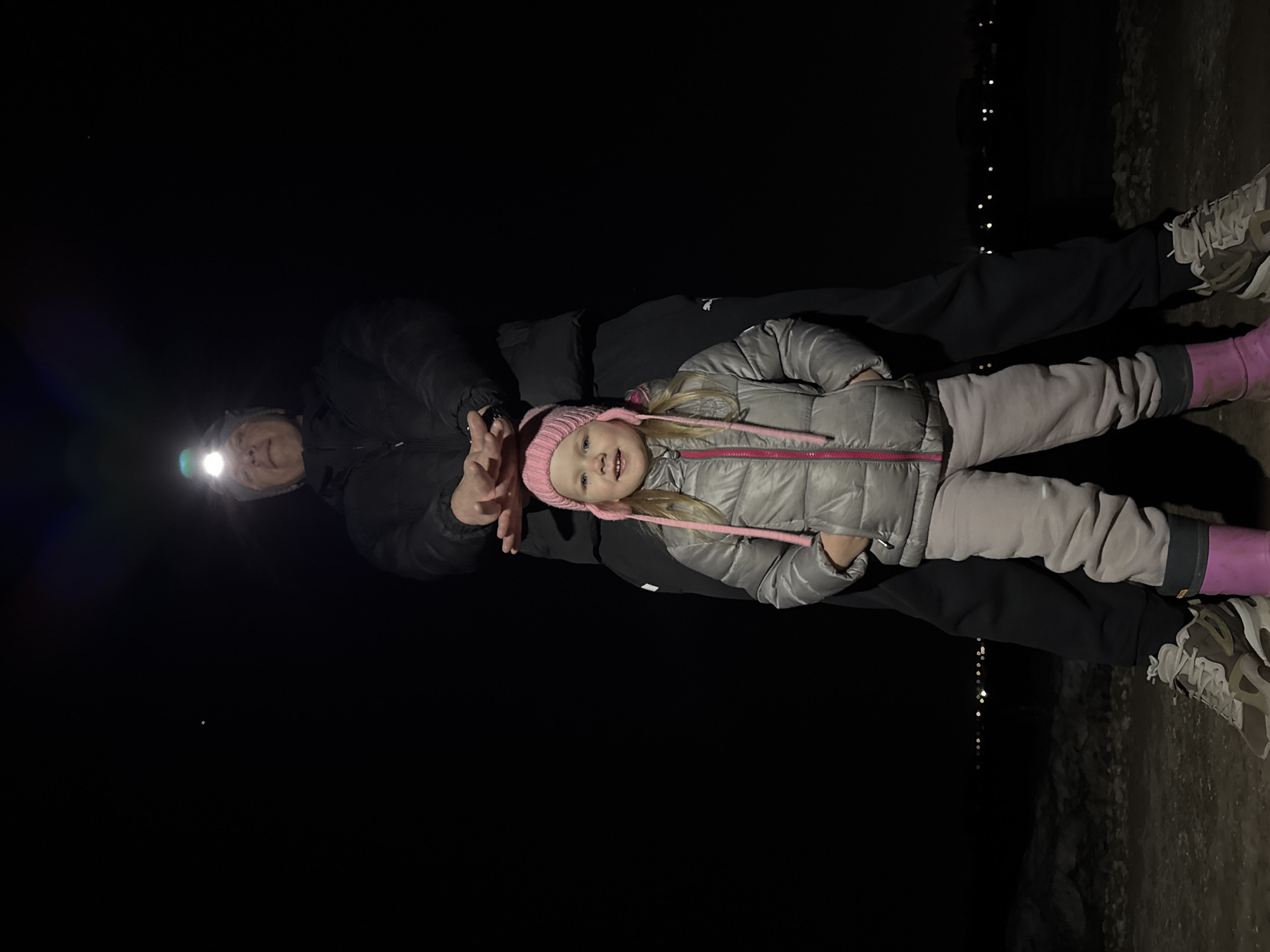 The image shows a young girl standing with an adult at night. The girl is wearing a pink hat, a silver jacket, grey pants, and pink boots. The adult is wearing a dark jacket and dark pants and has a headlamp on. The location is unclear but there are a few blurry lights in the distance and some grey pavement/ground under the feet of the subjects. It's not possible to determine if the location is Горный, Russia from the image alone.