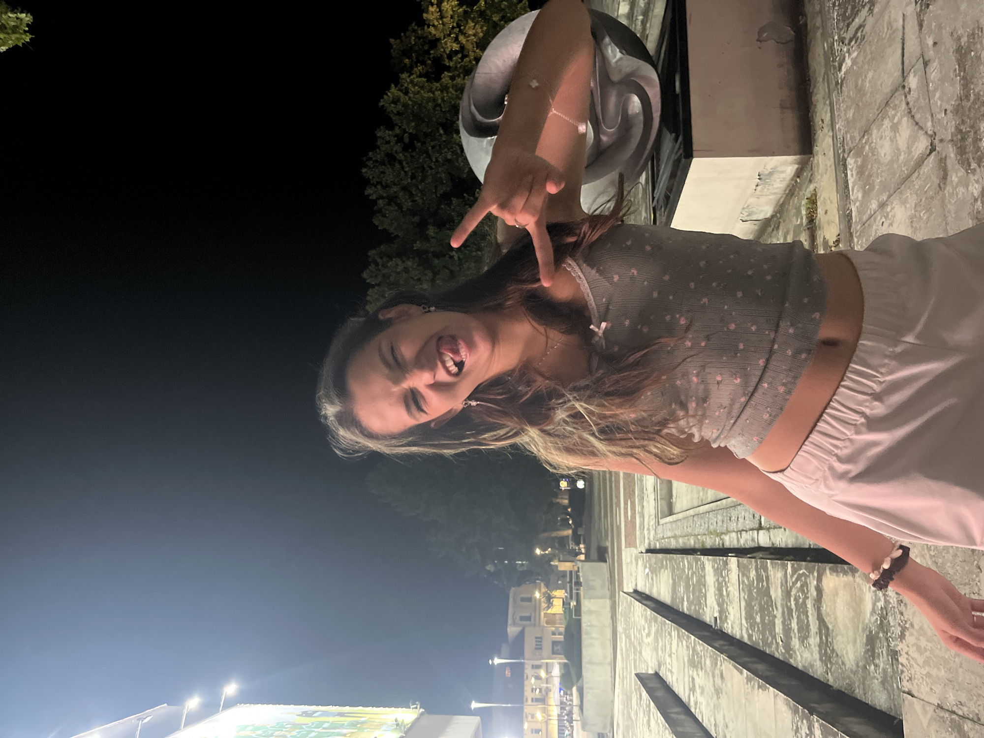 A young woman with long, wavy brown hair is pictured outdoors at night. She is wearing a gray crop top with a small floral pattern and light pink shorts. She is sticking out her tongue and making a "rock on" hand gesture with her right hand. Her left arm is bent and her hand is resting near her head. A bracelet with beads is visible on her right wrist.
The background suggests an urban setting at night. Trees are visible in the distance, as well as buildings with lights on. There appear to be streetlights and some illuminated signage. The ground in the foreground is a textured, concrete-like surface. The overall lighting indicates it is nighttime, with artificial lights illuminating the scene. The woman's expression is playful and energetic. The image appears to be a candid or posed photograph taken in a public space.