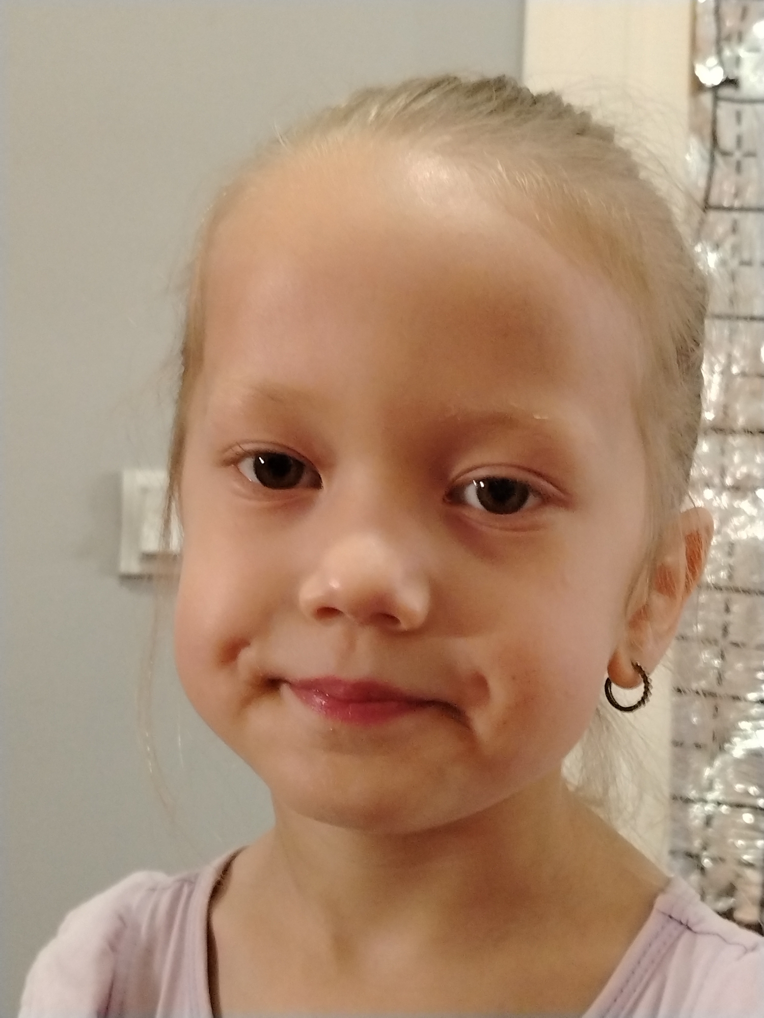 A close-up headshot captures a young Caucasian girl with light brown hair pulled back, facing forward. She has brown eyes, rosy cheeks with dimples, and slightly parted lips with a hint of lip gloss. She is wearing a light purple shirt and a small hoop earring in her right ear. The background is a light grey wall with a faint pattern on the right side. The lighting is soft and even. The image does not provide any information to indicate the location is in Shovot, Uzbekistan.