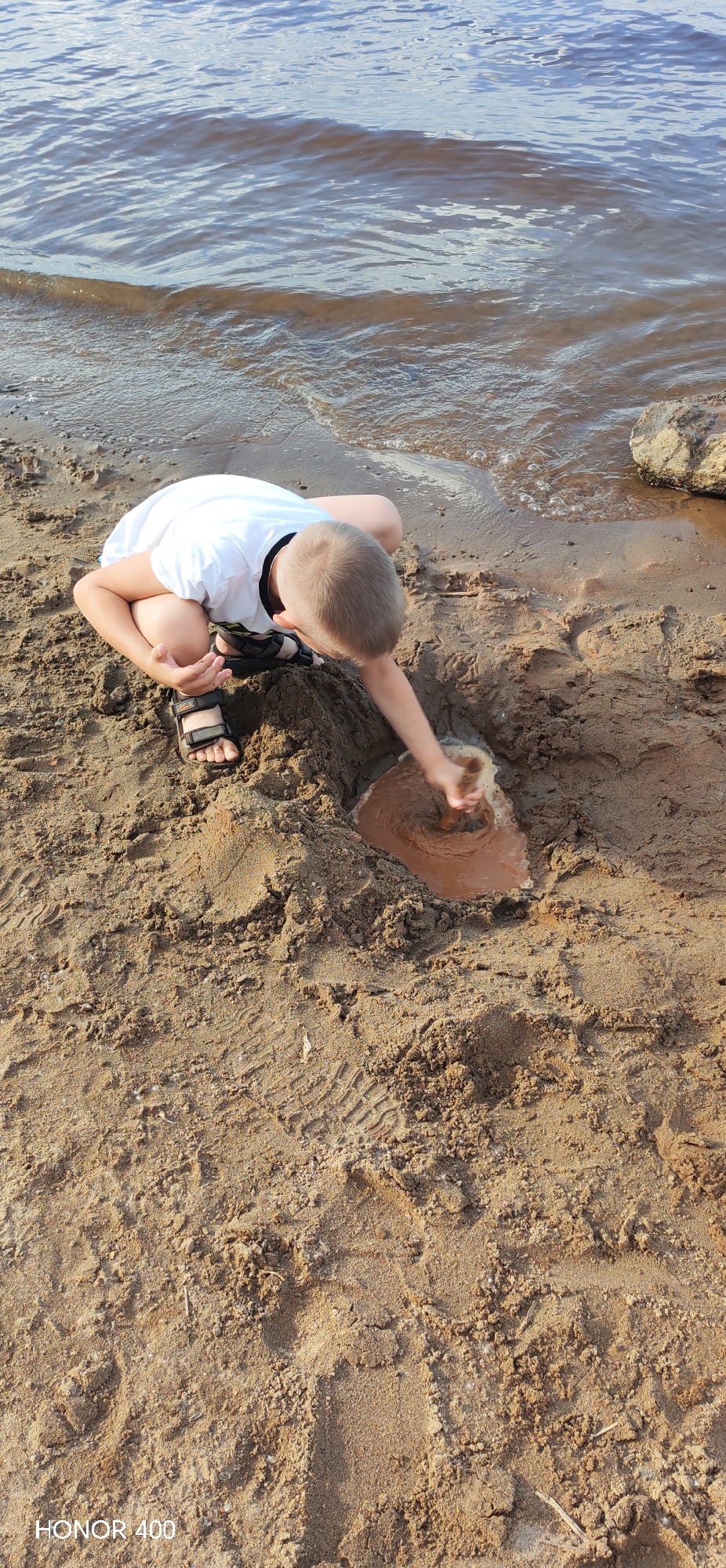A young boy with blond hair wearing a white t-shirt and dark sandals is crouched on a sandy beach near the Kama River in Chaikovsky, Russia. He is actively digging in the wet sand with his right hand, creating a swirling vortex of mud and water. The river water laps at the edge of the sandy shore, and a large rock is visible on the right side of the frame. The sand shows footprints, indicating previous activity on the beach. The image is captured from a high angle, looking down at the boy and his activity.