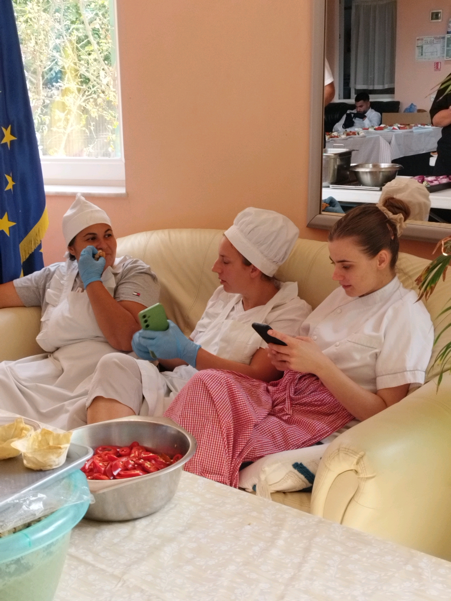 Three women, wearing white chef hats and aprons, are sitting on a light-colored sofa. The woman on the left is wearing blue gloves and holding a green smartphone. The woman in the middle is also wearing blue gloves and looking at her phone. The woman on the right is wearing a white chef jacket and looking at her phone. In the foreground, there is a bowl of sliced red peppers and some small pastry shells. The background shows a window with trees outside and an EU flag. A mirror reflects a man sitting at a table in the background, also looking at his phone. The scene appears to be indoors, possibly in a kitchen or dining area. The image does not provide any specific details to determine the location within Bucharest, Romania.