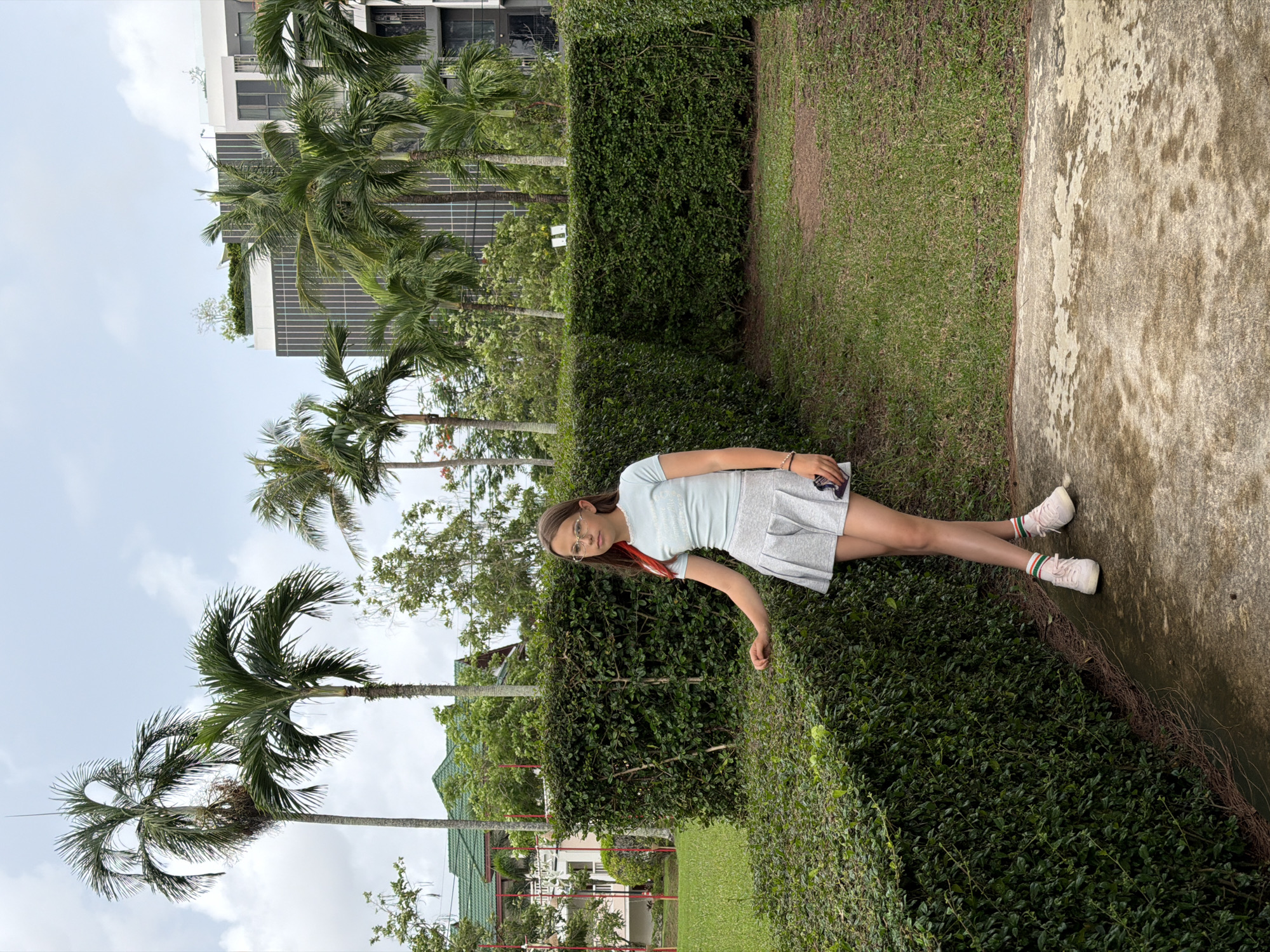 A young girl wearing a light blue t-shirt and a grey pleated skirt stands in a garden. She has on white sneakers with pink and green striped socks. She is wearing glasses and has a red ribbon in her hair. The girl is leaning against a tall, manicured green hedge. Behind her, there is a well-kept lawn, more hedges, and palm trees. In the background, a modern building with balconies and trees can be seen. The sky is overcast with clouds. The location appears to be a residential or resort area in Choeng Thale, Thailand, likely a common outdoor space within a condominium or housing development.