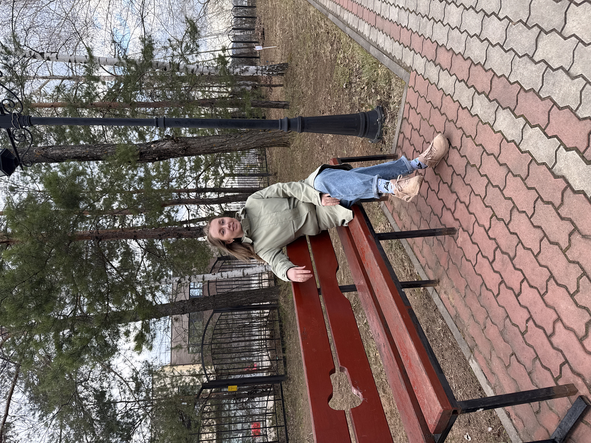 The image depicts a young woman lying on a red park bench. She is wearing a light green jacket, blue jeans, and tan shoes. The bench is situated next to a patterned brick walkway. A black lamppost stands to the left of the bench. Trees are visible in the background behind a black iron fence. The location is likely a park in an urban setting.