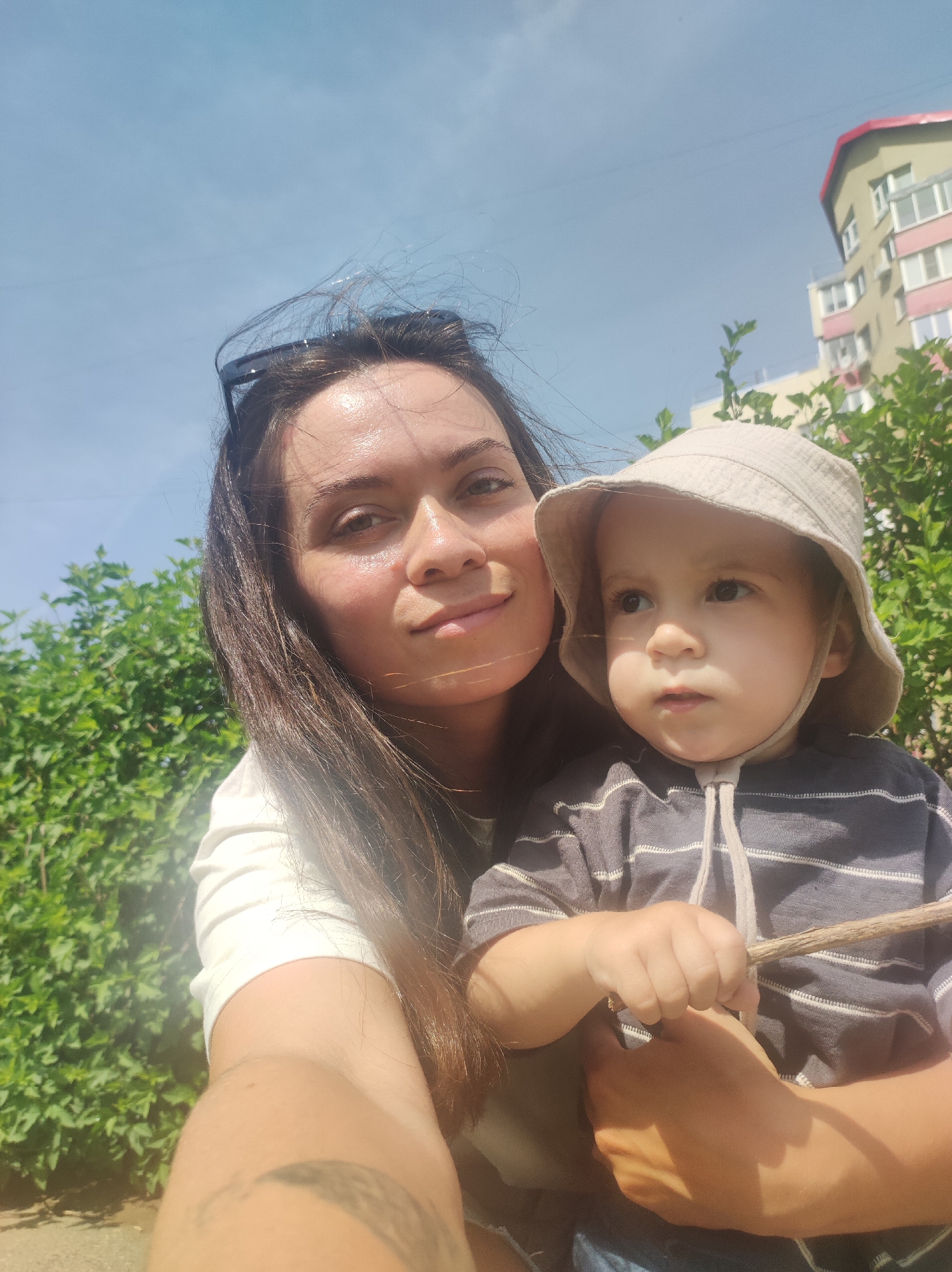 The image shows a selfie of a woman holding a young child. The woman has long, dark hair, is wearing sunglasses on top of her head, and has a white top. The child is wearing a beige bucket hat and a striped grey and white shirt, and is holding a small stick. In the background there is a green bush, a building with white and pink elements, and a blue sky. The location is Chaikovsky, Russia, judging by the building and general appearance.