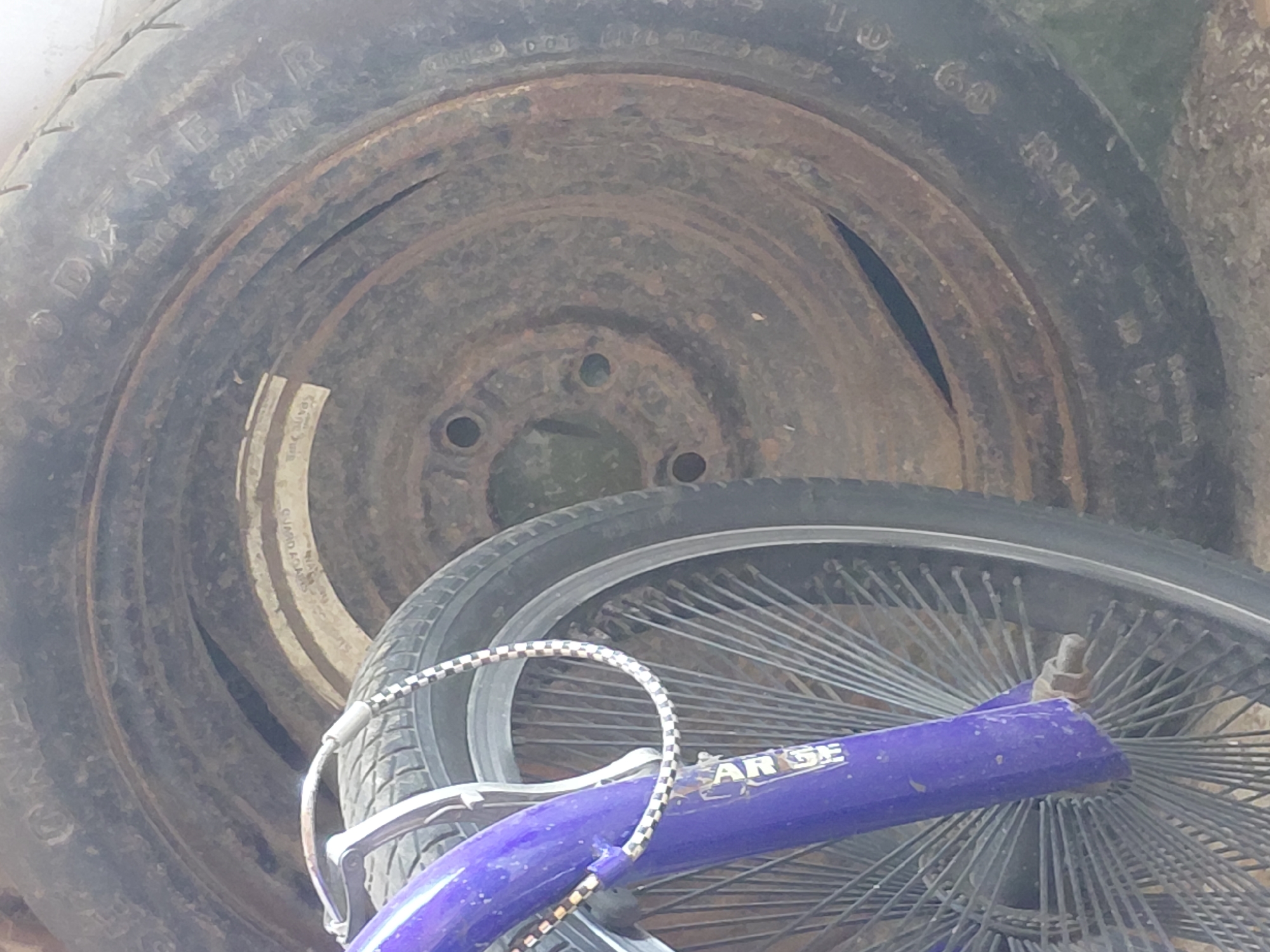 The image shows a close-up view of a bicycle wheel and a spare tire, likely in an outdoor setting. The bicycle wheel, with its numerous thin spokes, is partially visible on the left side of the frame and is colored purple. A checkered cord is wrapped around the handlebars. To the right of the bicycle wheel, a spare tire is positioned. The tire appears old and has markings that are partially obscured. The center of the tire is visible, revealing a rusty metal rim with three circular holes. The overall scene suggests a storage area or a less-maintained environment. No specific details about Santa Ana, Venezuela are discernible from the image itself.