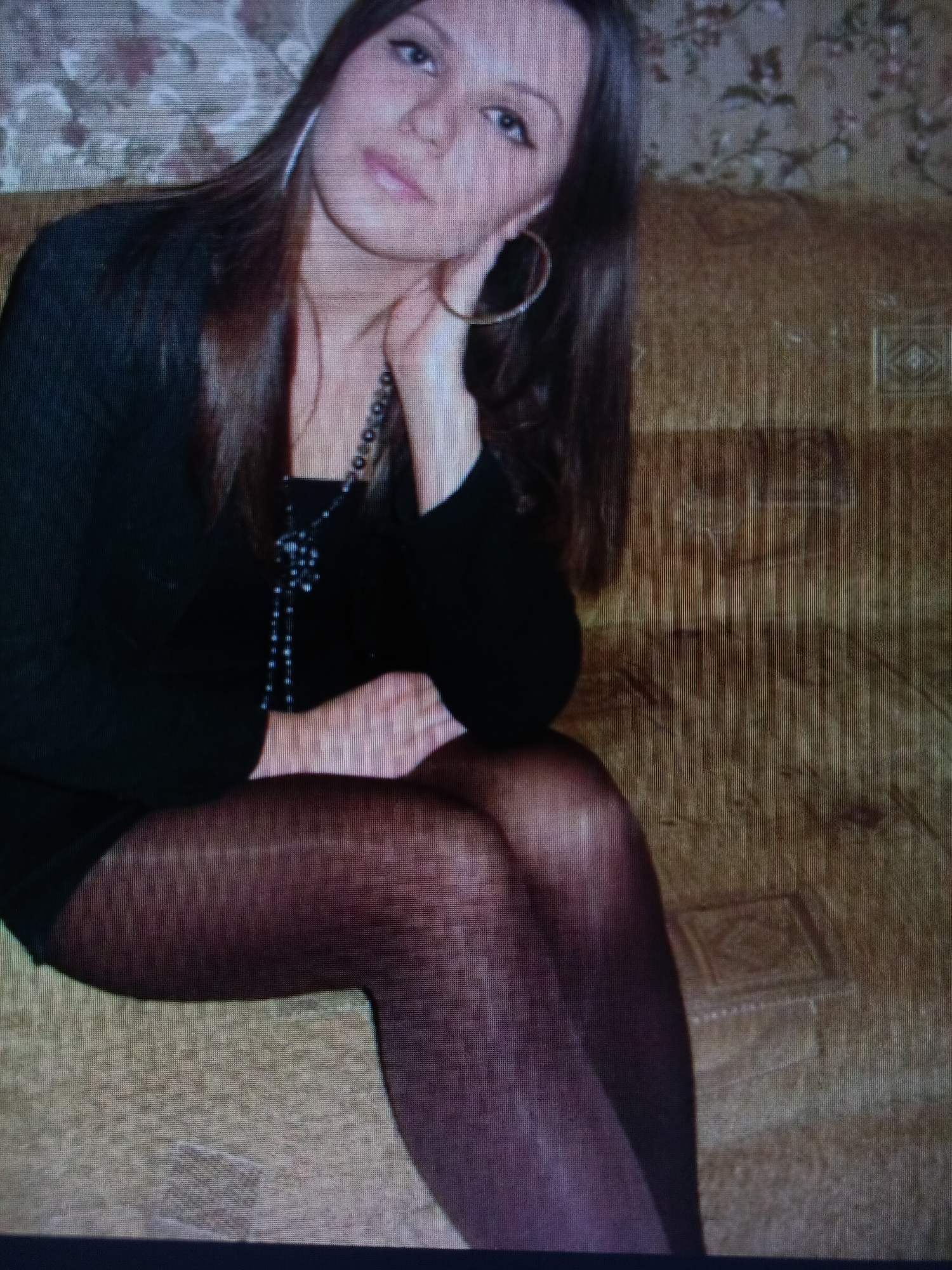 A young woman with long brown hair and large hoop earrings is sitting on a textured brown couch. She is wearing a black top and dark pantyhose. She has a necklace with dark beads. The background is a floral patterned wallpaper. The image is of low quality and appears to be taken with a digital camera. The location is not specified in the image and cannot be determined.