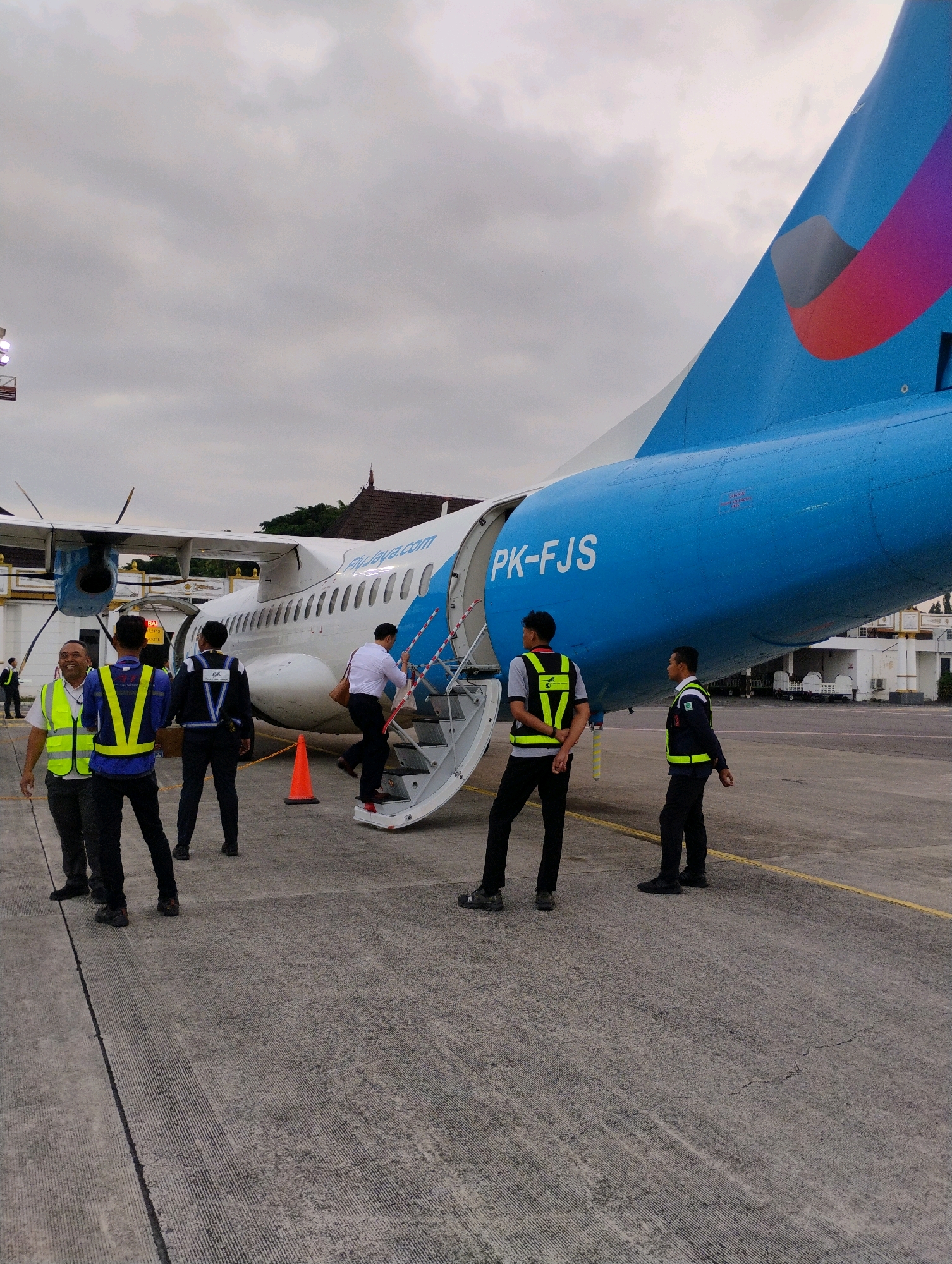 An overcast day in Maguwoharjo, Indonesia, where a bright blue and white airplane with the registration "PK-FJS" is parked on the tarmac. Several individuals in high-visibility vests, likely airport ground crew, are present near the open boarding stairs. One person is ascending the stairs, dressed in a white shirt and dark pants, carrying a bag. The tail of the plane features a colorful logo in shades of pink, purple, and gray. The scene appears to be an active airport environment.