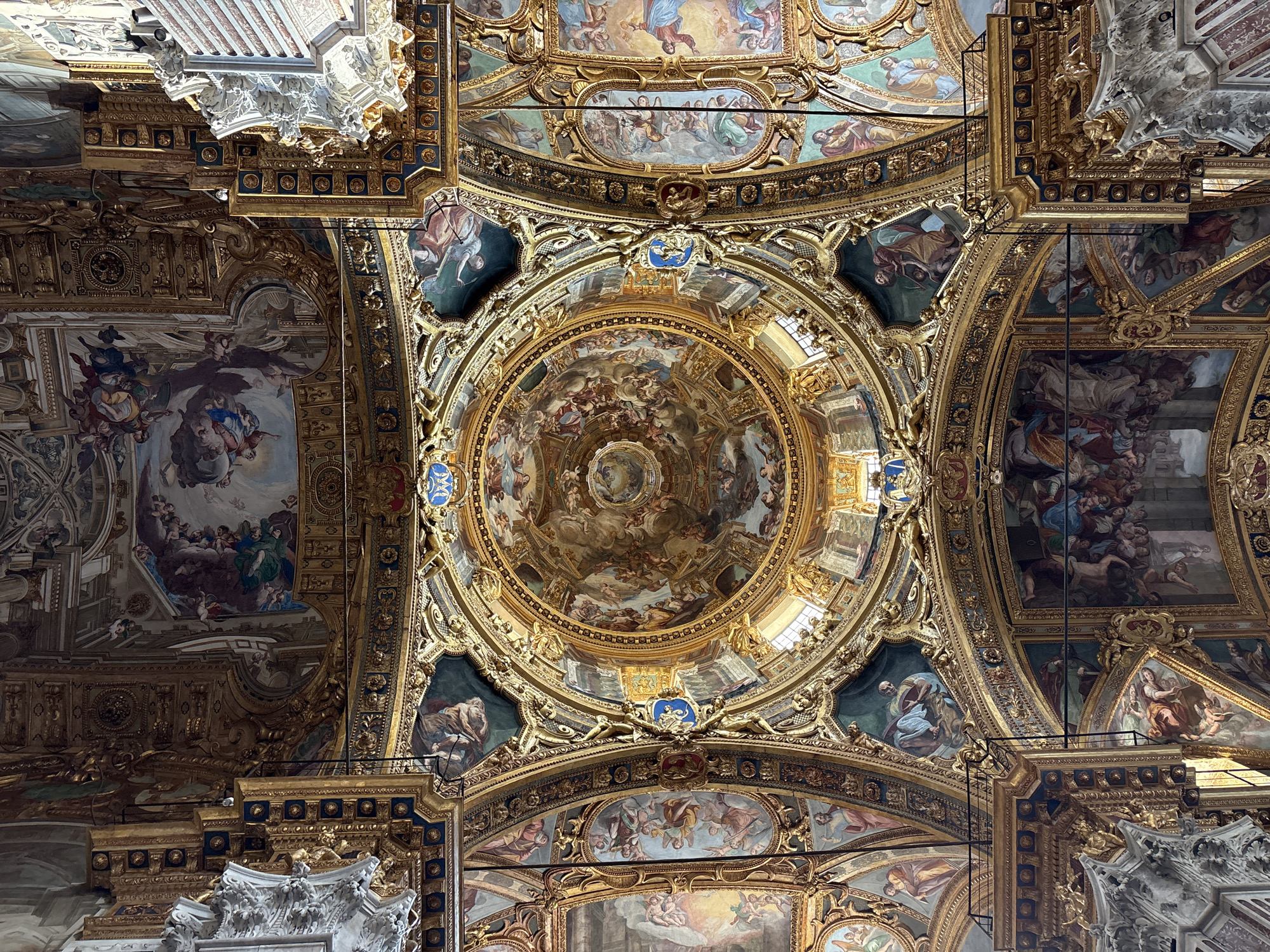 This media file captures an opulent, highly ornate ceiling, typical of Baroque or Rococo architecture, likely within a grand church or palace in Genoa, Italy. The view is directly upwards, emphasizing the intricate design and vastness of the decorated surface.
The central feature is a large, circular dome adorned with a vibrant fresco. This fresco depicts a multitude of figures – people, angels, and cherubs – seemingly ascending or gathered in a celestial realm amidst clouds, with light radiating from its center, creating a sense of spiritual awe and movement.
Surrounding the central dome are vaulted sections and numerous rectangular and oval panels, each containing separate painted scenes. These scenes also feature various human figures and angels, suggesting religious narratives or allegorical representations, all executed in rich, warm colors.
The painted sections are elaborately framed by extensive three-dimensional gilded stucco work. This intricate gold ornamentation features scrolling patterns, acanthus leaves, classical motifs, and small sculptural elements like putti or cherubs. Sections of deep blue accent the gold, providing a striking contrast to the vibrant colors of the frescoes. Small blue shields or cartouches, some bearing golden crest-like symbols, are strategically placed within the gilded framework.
The lighting appears bright, likely from natural daylight entering through unseen windows, illuminating the intricate details and rich colors. Minimal modern elements, such as small black fixtures (possibly lights or sensors), are sparingly visible, contrasting with the historical artistry. No discernible text is visible within the image. The overall impression is one of immense grandeur, artistry, and religious or historical significance.