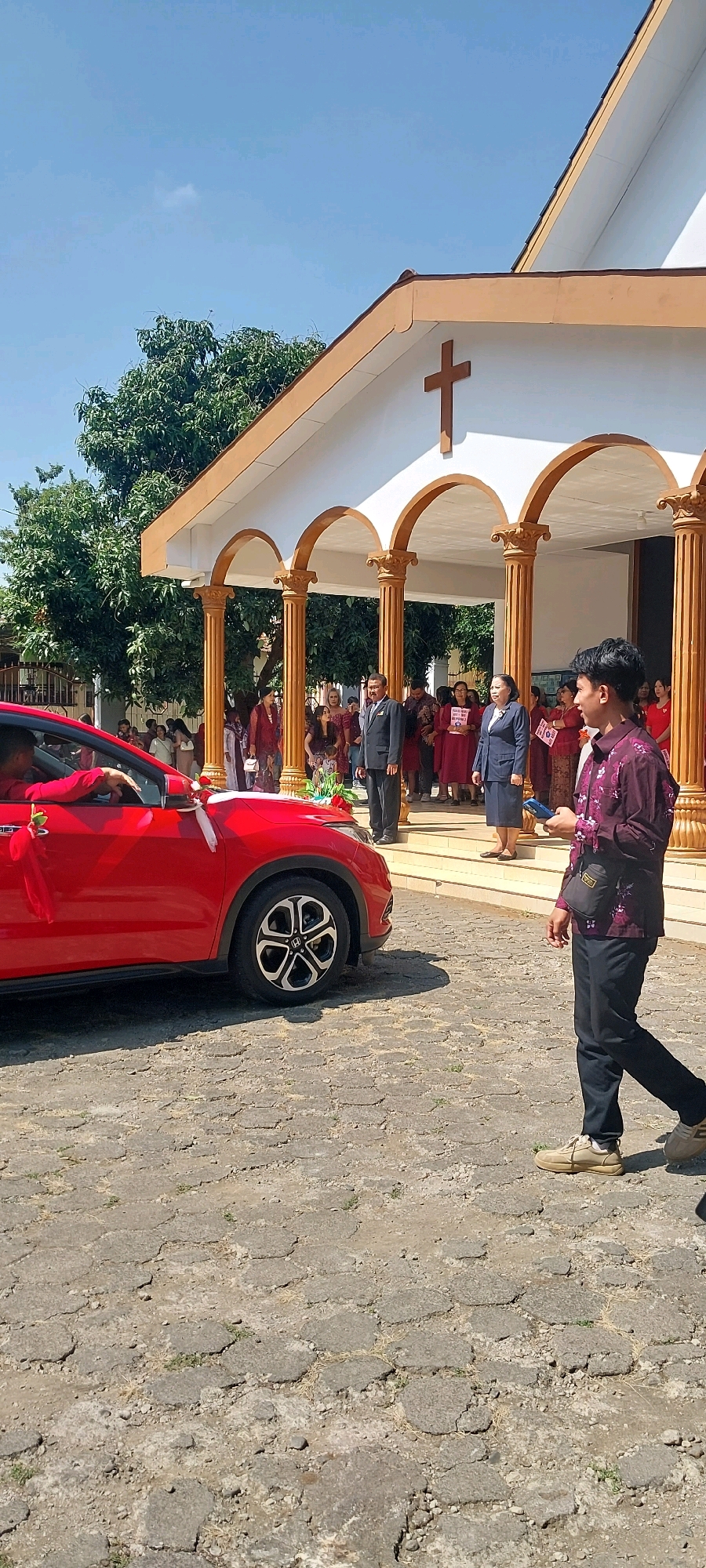 An outdoor scene in front of a church in Keling, Indonesia, under a clear blue sky. The church has a white facade with a prominent brown cross at the apex of its gable. A covered entryway is supported by golden Corinthian columns. To the left, a red car is parked, decorated with white and red ribbons and a rose. A person in a red shirt is visible inside the car. In the foreground, a young man wearing a maroon patterned shirt, dark pants, and light-colored shoes walks towards the right, holding a blue object, possibly a phone, and carrying a black satchel. A group of people, some in traditional attire, are gathered in front of the church. Two individuals in formal wear, a man in a dark suit and a woman in a dark suit, stand near the church entrance, facing the gathered people. The ground is paved with irregular, hexagonal stones. Lush green trees are visible in the background.