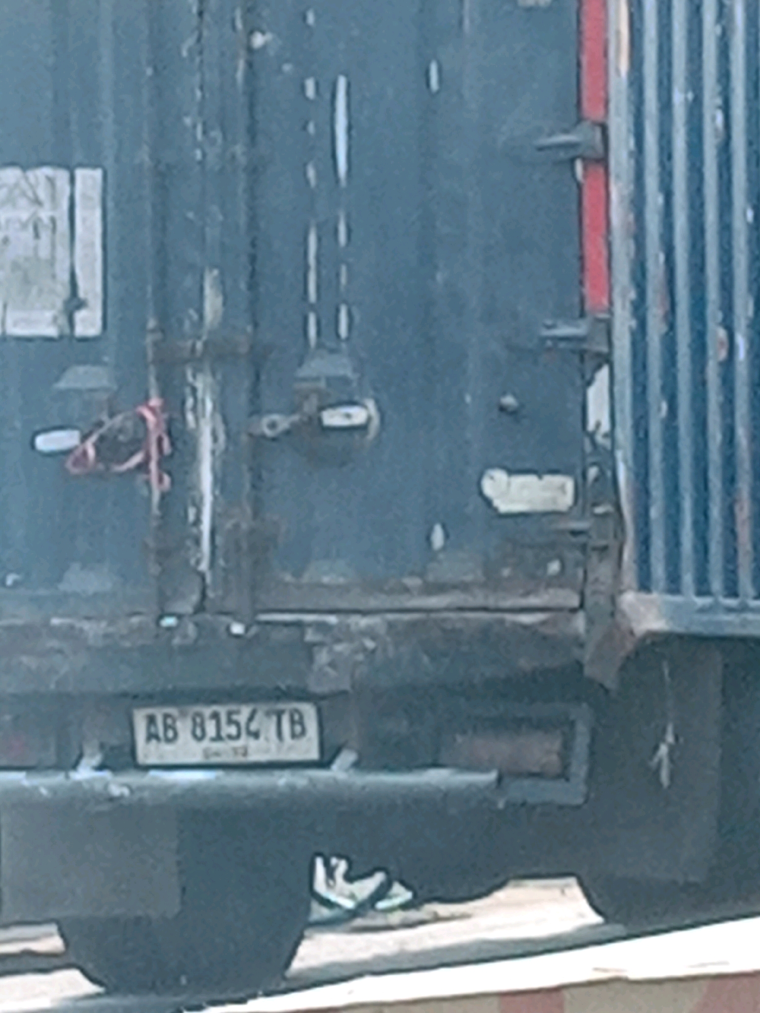 The image captures a close-up view of the rear of a dark-colored vehicle, possibly a shipping container or truck, in Banguntapan, Indonesia. A license plate with the characters "AB 8154 TB" is visible, indicating its registration. The vehicle's surface shows signs of wear and tear, with rust and chipped paint. There are several metal fixtures and a red strap attached to the rear door mechanism. In the lower portion of the image, a pair of white sneakers is partially visible, suggesting the presence of a person or people nearby, though they are not clearly depicted. The ground appears to be a paved surface, likely a road or industrial area.