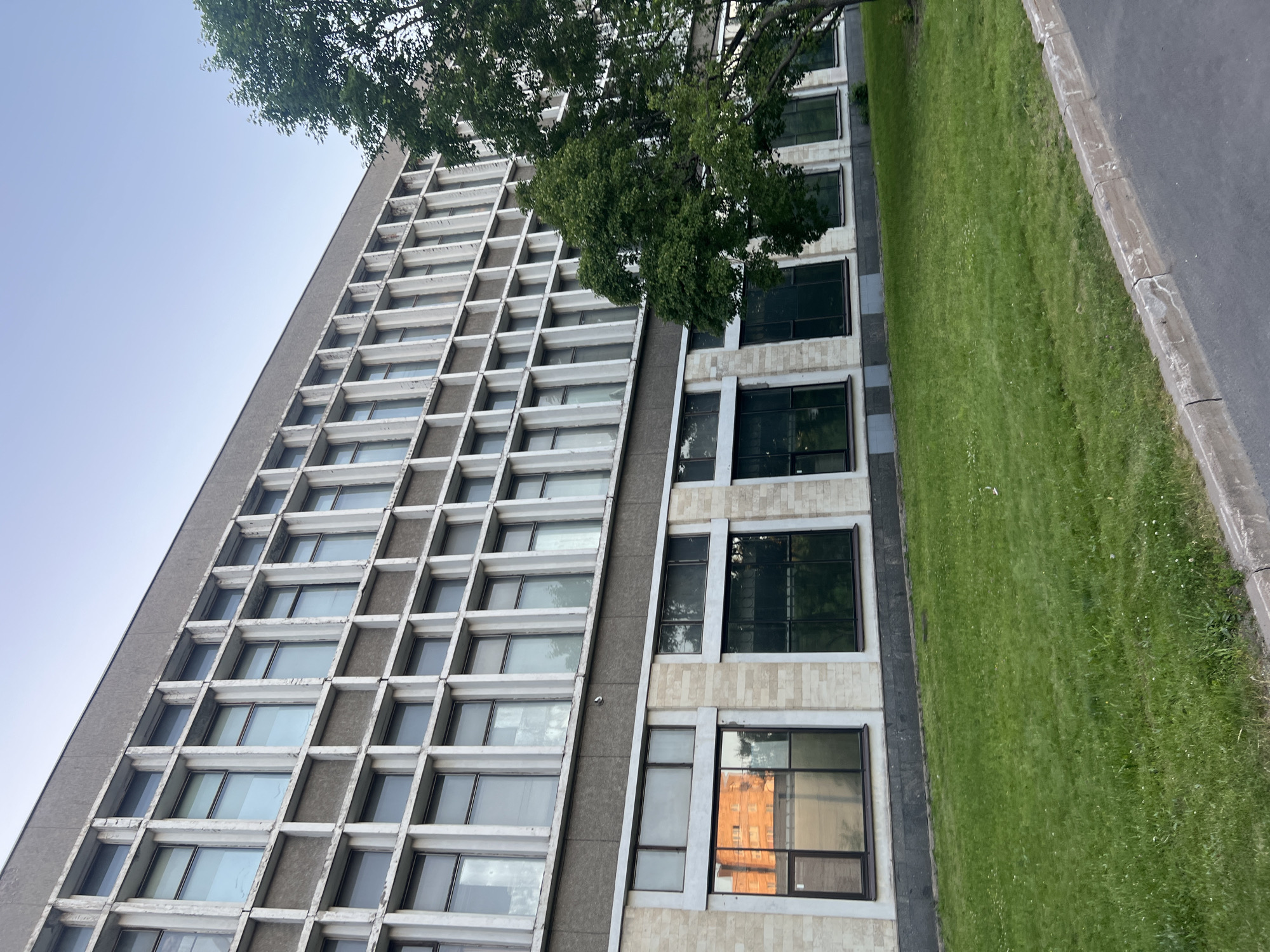 An institutional-style building in the city of Unknown, Russia, is depicted under a clear, light blue sky. The multi-story structure features a grid-like facade on its upper section, composed of numerous vertically oriented rectangular windows set within light-colored concrete or stucco frames. The grey-brown panels between these windows show signs of wear, discoloration, and minor material degradation. The windows reflect the sky and ambient light. A darker, horizontal band separates these upper floors from a lower section, which presents larger, horizontally oriented windows with dark frames, set within a lighter, possibly stone or textured stucco facade. One lower-level window prominently reflects an orange-hued building. A mature deciduous tree with dense green foliage partially obscures the right side of the building. In the foreground, a vibrant green lawn, dotted with small white flowers, slopes gently towards a concrete curb. Beyond the curb, a dark asphalt or paved surface is visible. No individuals or animals are present in the scene.