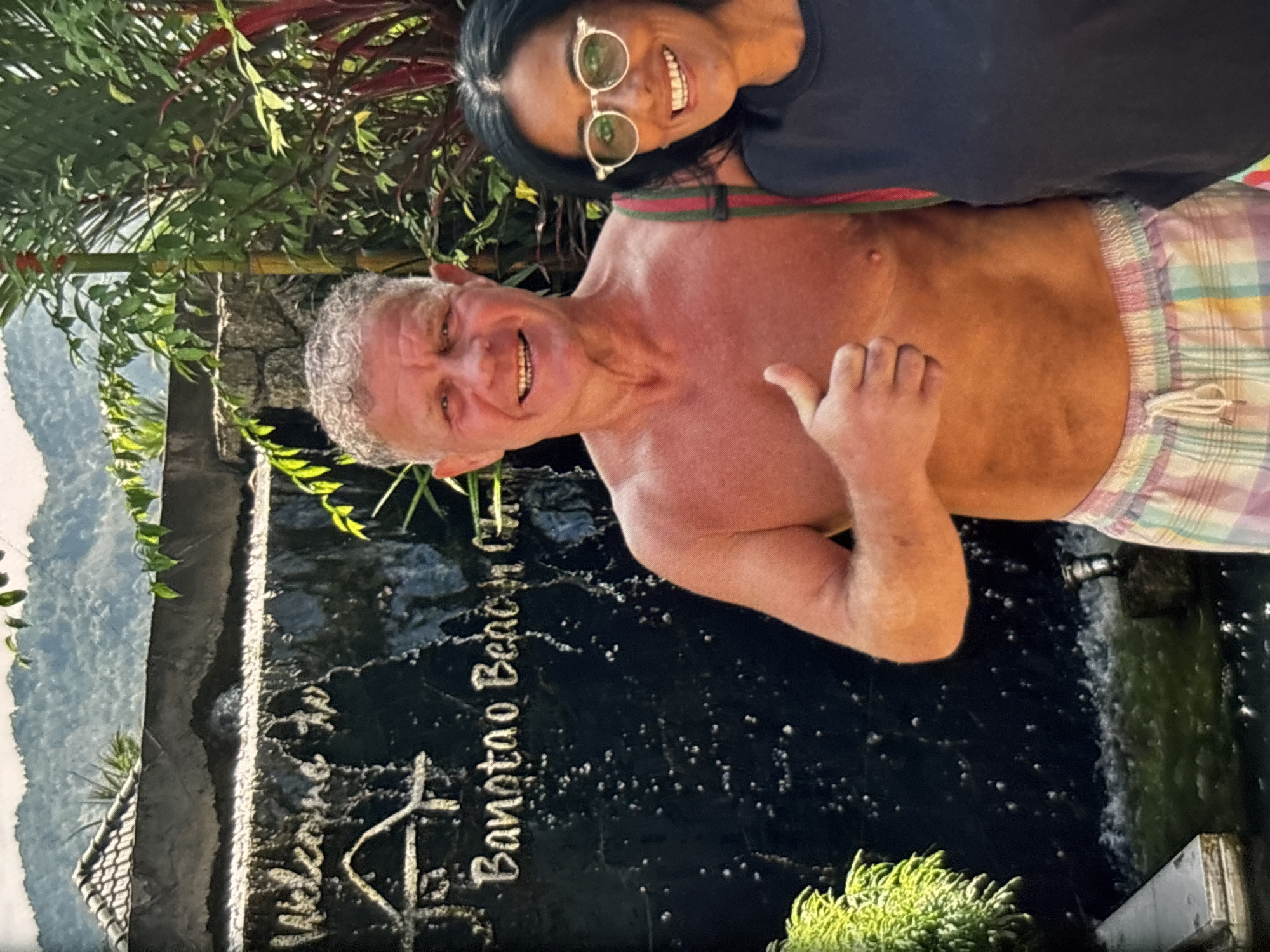 A shirtless man with grey curly hair and a woman wearing glasses and a black shirt are standing in front of a waterfall with a sign that reads "Welcome to Banotoa Beach". The man is giving a thumbs up. The location appears to be outdoors with lush greenery and mountains in the background, consistent with a tropical setting. The man is wearing plaid shorts, and the woman has a striped strap visible on her shoulder. The image quality is somewhat low, with some pixelation and a grainy appearance. The lighting suggests it is daytime.