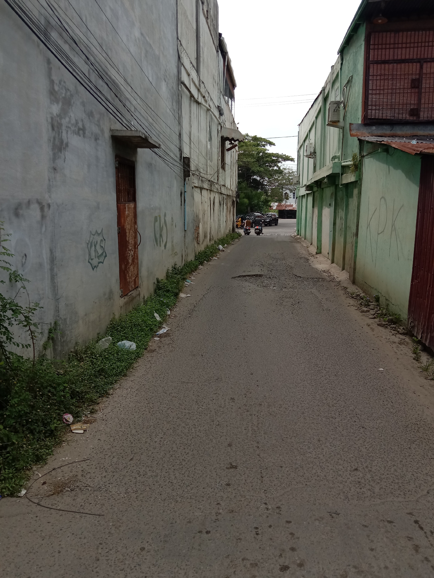 The image depicts a narrow, poorly maintained alleyway in Lhokseumawe, Indonesia. The road surface is cracked and potholed. Buildings flank both sides, their walls displaying weathered paint, graffiti, and exposed utility lines. Vegetation and litter are visible along the edges of the road. In the distance, motorbikes and parked cars suggest moderate traffic. The sky is overcast.