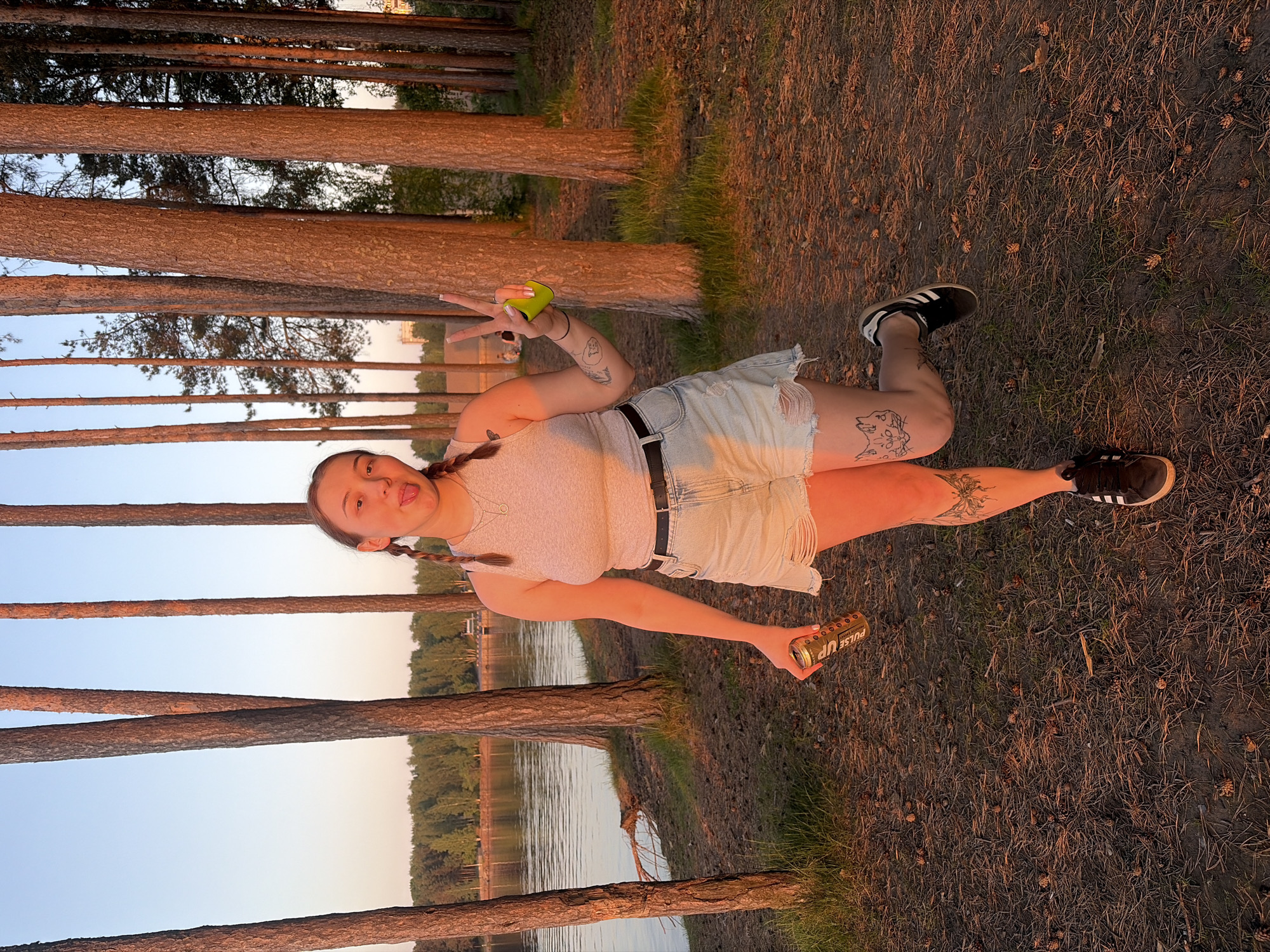 A young woman lies on the ground in a pine forest near a body of water. She is wearing a light-colored tank top, denim shorts with a black belt, and sneakers. She is holding a green object in one hand and a can of "Pulse Up" energy drink in the other. She is sticking her tongue out and making a peace sign with her fingers. Tattoos are visible on her arms and legs. The ground is covered in pine needles and small pine cones. Tall pine trees surround the scene, with a lake and forest visible in the background. The sky is light blue, suggesting it is dusk or dawn. The scene is lit with a warm, golden light. The location is in the city of Chaikovsky, Russia.
