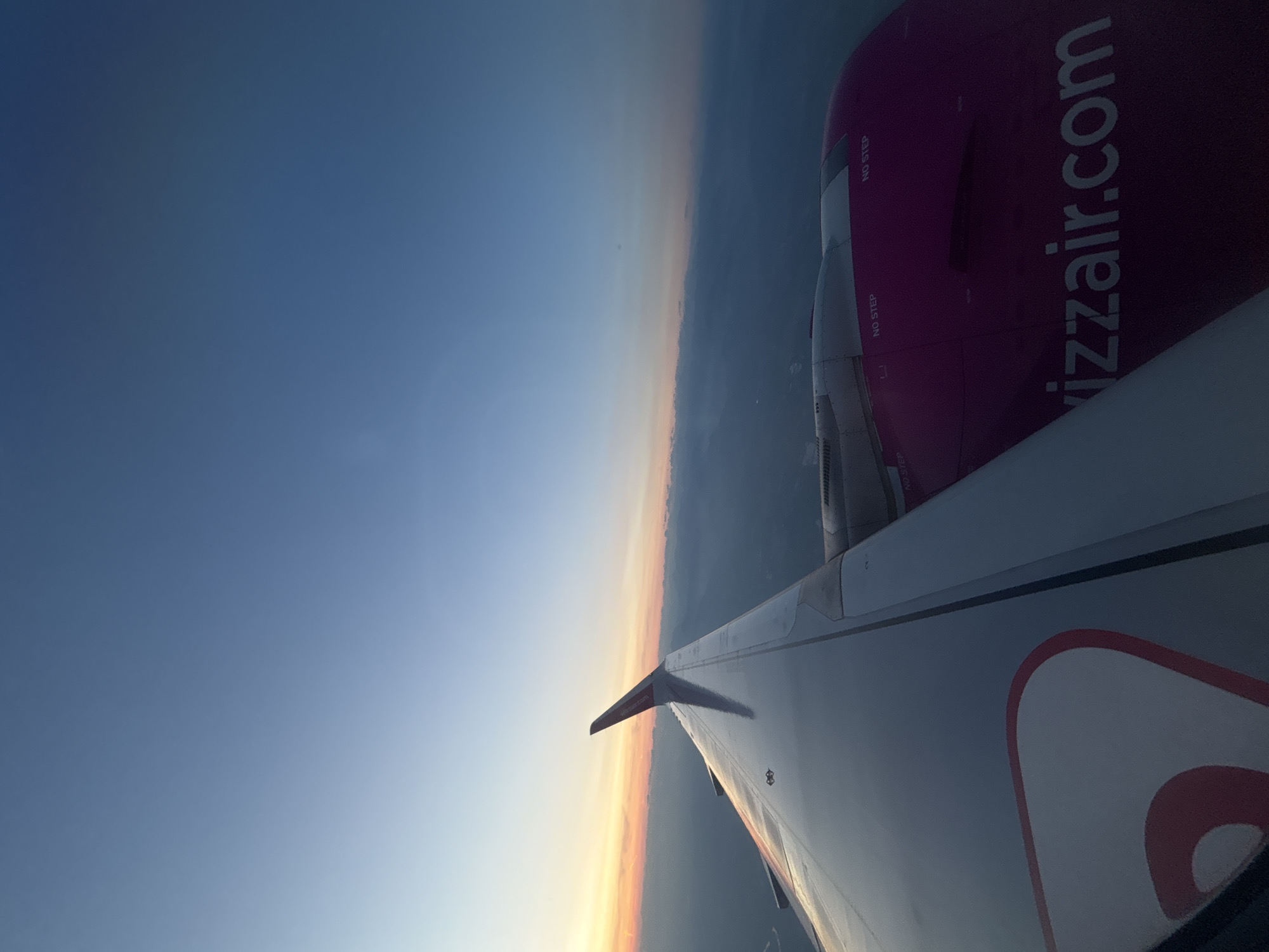 This is an aerial photograph of the wing and engine of a Wizz Air airplane, seen from a passenger's perspective during flight. The wing is predominantly silver with a red and white design on its surface, and it extends diagonally from the lower left corner towards the center of the frame. The engine, painted in a vibrant magenta color, is located towards the right side of the image and prominently displays "wizzair.com" in white lettering. Two "NO STEP" markings are visible on the engine.
The background consists of a twilight sky transitioning from a pale orange and yellow horizon, where the sun is likely setting or rising, to a deep blue overhead. Below the horizon, a layer of clouds or haze partially obscures the landscape, which appears to be rural or semi-urban. The image captures the vastness of the sky and the perspective of being high above the ground. There are no people visible in the image. The location in the city of Pagazzano, Italy is not discernible from the visual information provided in the photograph.