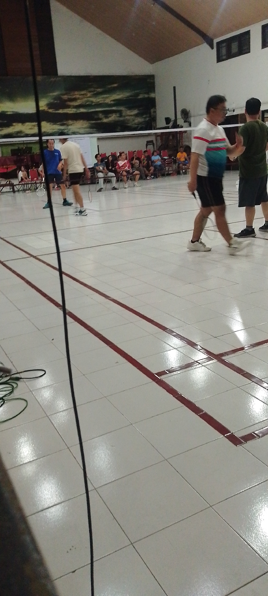 This image depicts an indoor badminton court with tiled flooring marked with red lines. In the foreground, a black cable lies on the floor, extending from the lower left corner towards the center. Two men are visible in the middle ground, actively engaged in a badminton match. One man, wearing a white t-shirt with red and blue stripes and black shorts, is in motion. The other man, in a dark green t-shirt and black shorts, is also actively playing. In the background, spectators are seated on chairs, observing the game. The court is illuminated by overhead lighting, and a large mural is visible on the wall behind the net. Based on the context of similar images, this scene is likely within the Djarum Kudus Sports Hall in Kudus, Indonesia, a known venue for badminton activities.