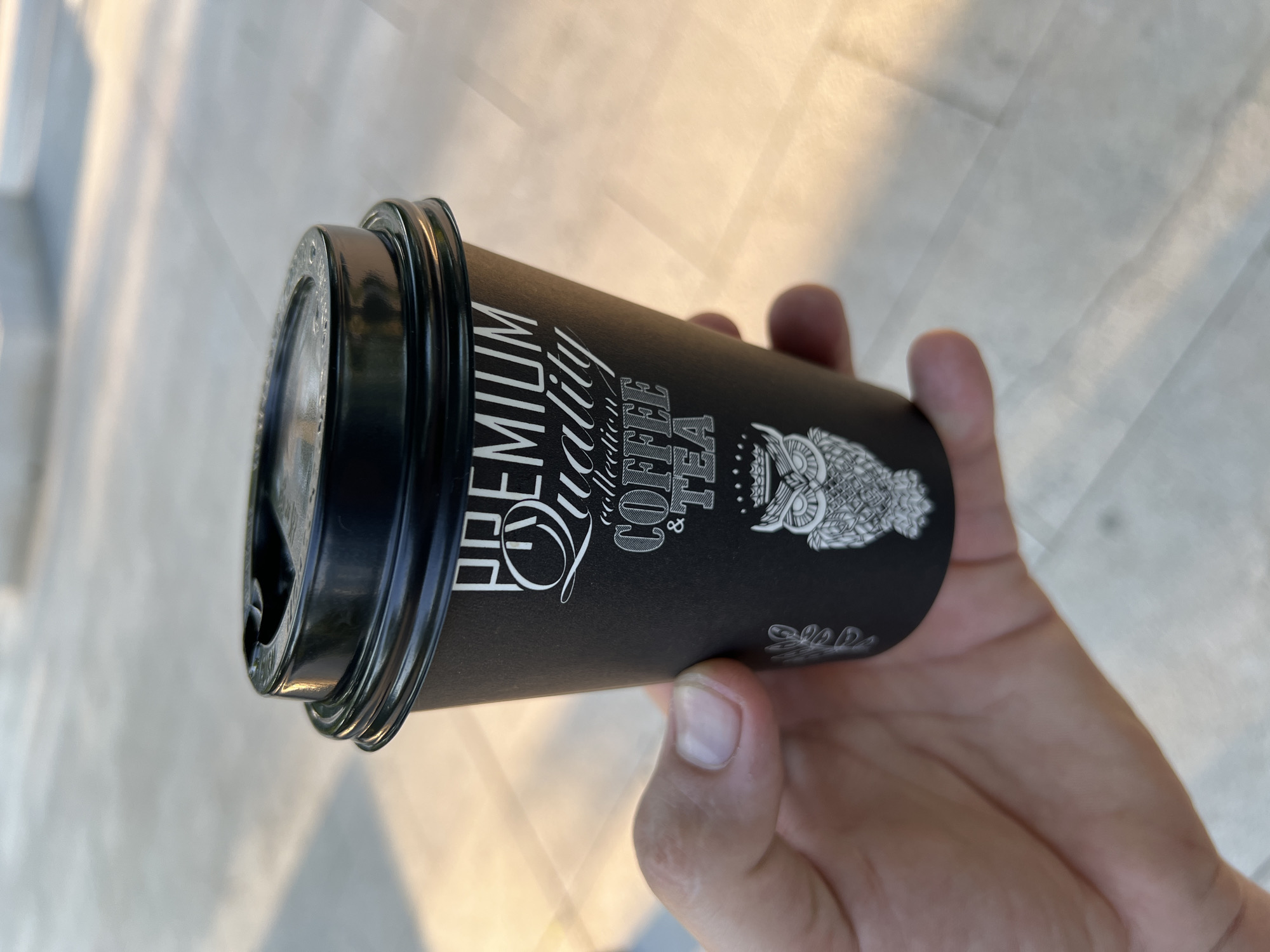 A person's hand holds a black paper coffee cup with a black plastic lid. The cup has white text on it that reads "PREMIUM quality, Coffee & Tea," and a stylized drawing of an owl. The scene takes place outside on a sunny day. It's located in Krasnodar, Russia.