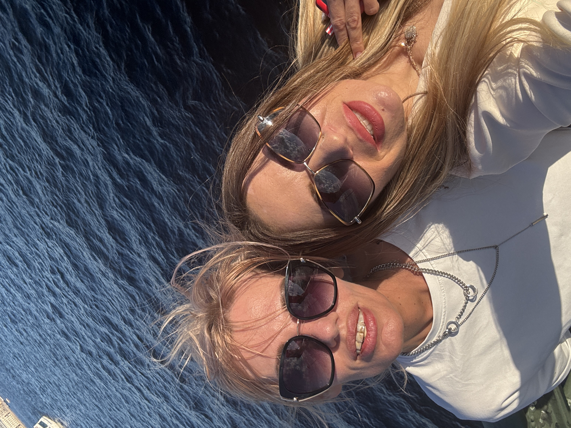 Two women, wearing sunglasses and smiling, pose for a selfie on a boat sailing on the Malaya Neva River in Saint Petersburg, Russia. The woman on the left, with blonde hair, wears a white t-shirt and a silver necklace. The woman on the right, with long blonde hair, wears a white top and a necklace with a clover pendant. In the background, the choppy, dark blue water of the river reflects the sunlight. The city skyline is visible in the distance on the left side of the image.