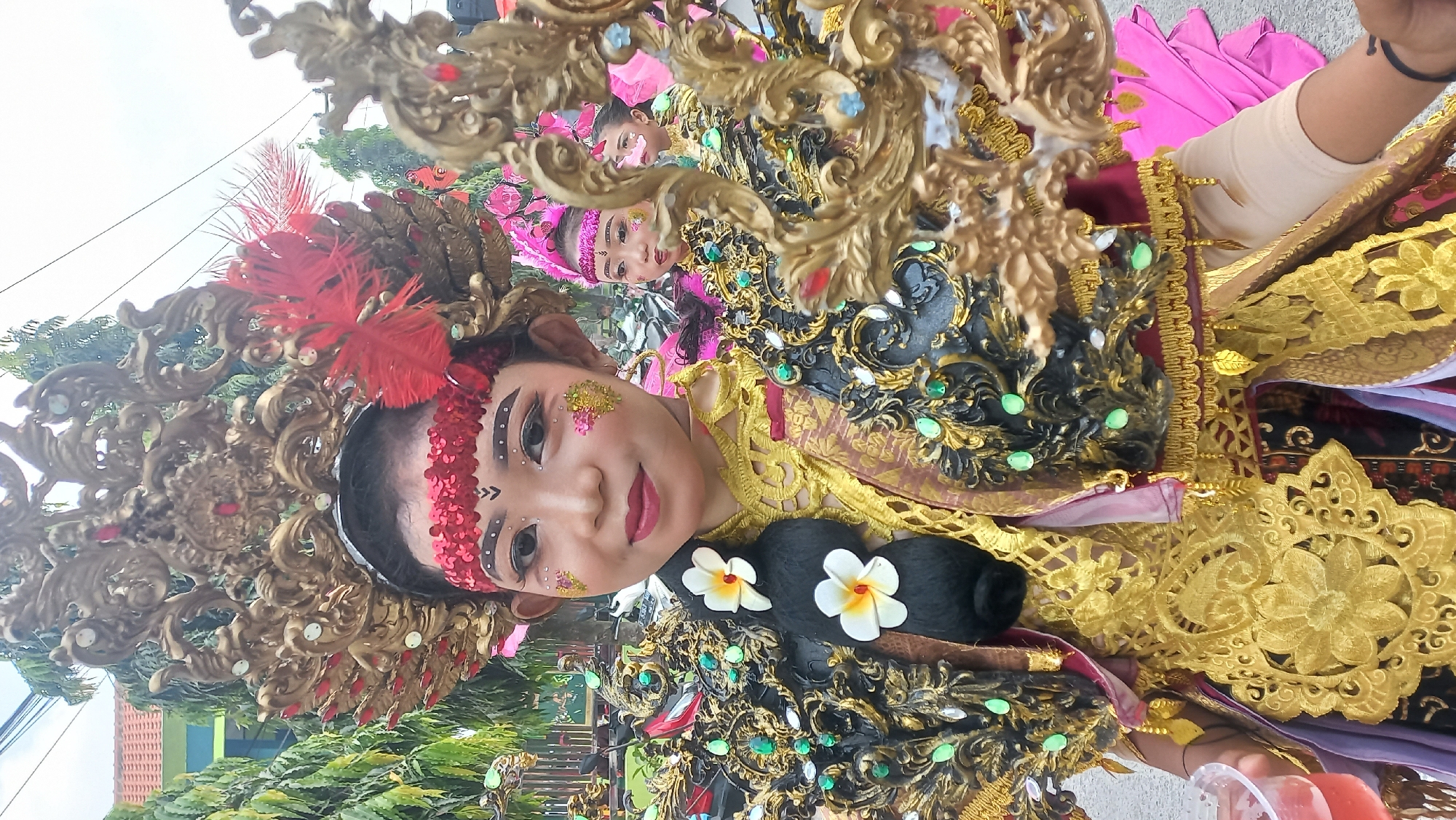 A close-up, outdoor shot captures a young girl in traditional attire for a parade in Kedungmegarih, Indonesia. The girl, positioned centrally and facing the camera, wears an elaborate headdress adorned with gold filigree and red feathers. Her face is painted with decorative makeup, including red lipstick, white and black eyeshadow, and gold glitter accents on her cheeks. Her hair is styled in a braid adorned with two white flowers. She is dressed in a richly decorated costume featuring gold embroidery, black and green embellishments on the shoulders, and a golden ceremonial cloth around her neck. To the right of the main subject, other children in similar parade attire can be seen. The background suggests an urban setting with a building and trees, with power lines visible overhead. The ground appears to be asphalt.
