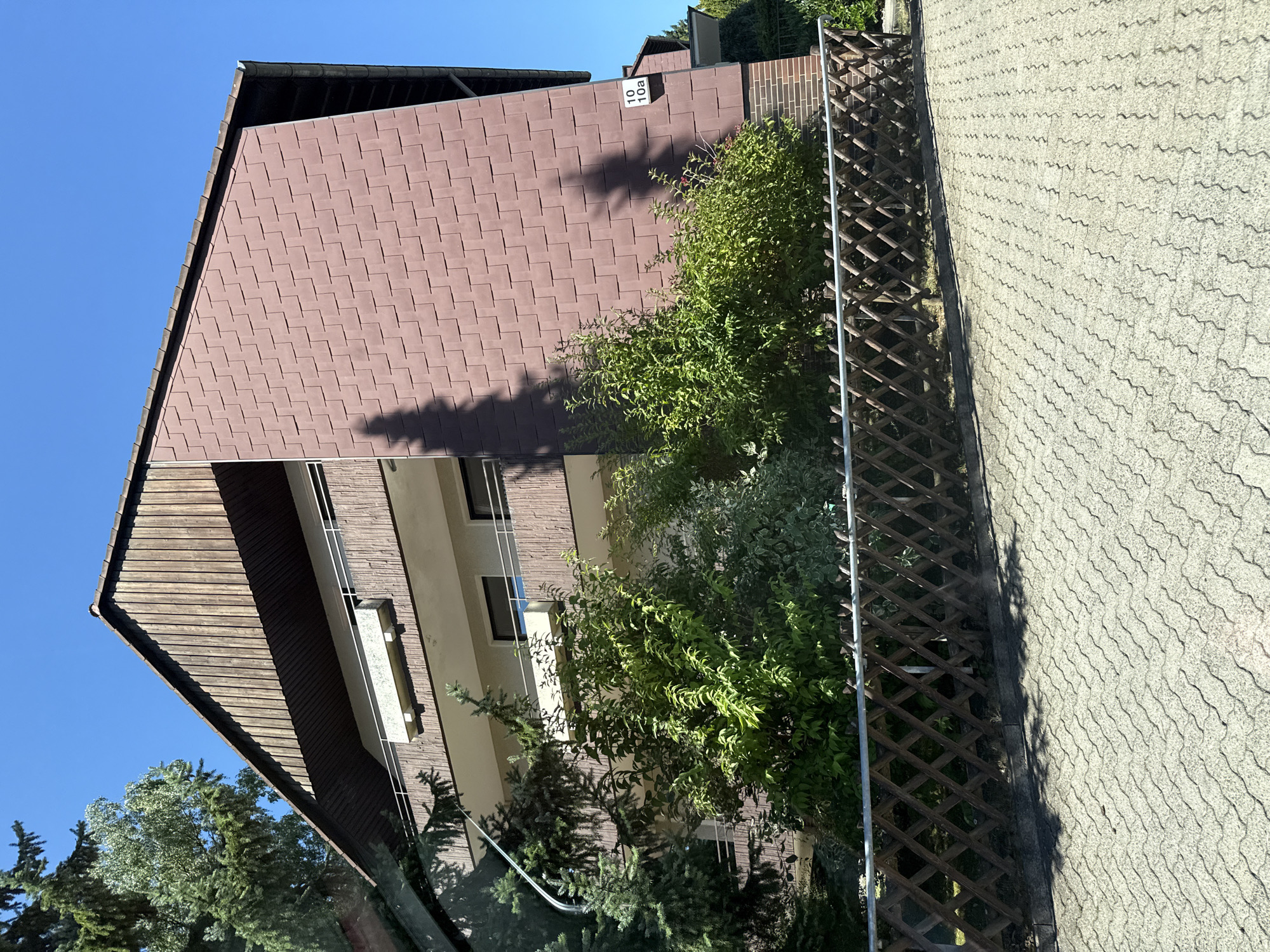 The image shows the exterior of a multi-story building with a brown, shingle-like facade on the upper portion and balconies. The building appears to be residential. A number sign on the facade reads "10 10a". Trees and bushes are visible in front of the building, partially obscuring the lower levels. A wooden lattice fence runs along the front of the property. The ground in front is paved with interlocking concrete blocks. The sky is clear and blue, indicating a sunny day. The scene is likely in a suburban or residential area of Bad Sachsa, Germany.