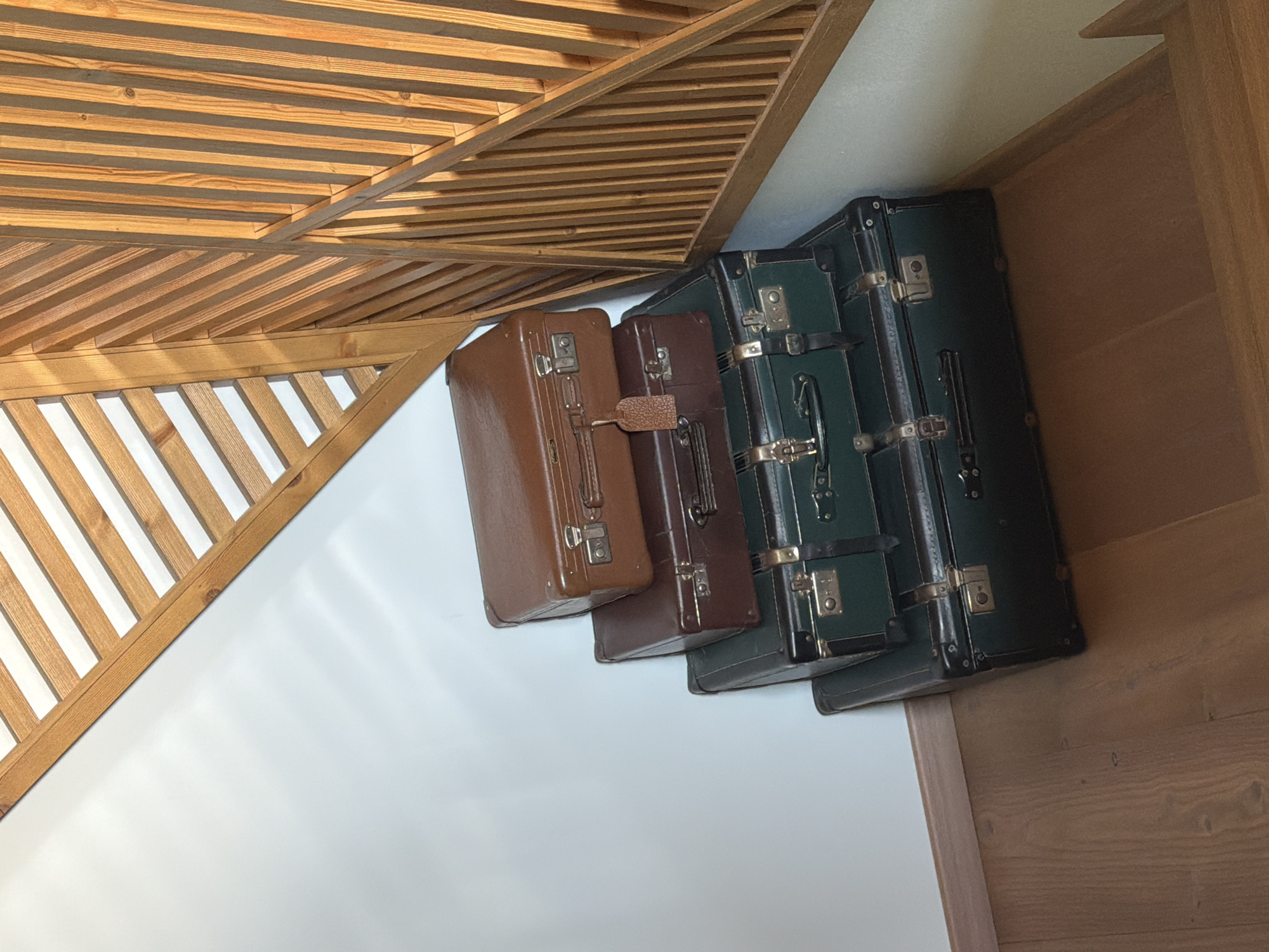 Four vintage suitcases are stacked against a white wall, with a wooden ceiling structure above and a wooden floor to the right. The suitcases are in varying shades of brown and green, with metal clasps and handles. The top suitcase is brown, followed by a dark brown one, and then two dark green suitcases. The lighting suggests it is daytime, with natural light casting shadows from the wooden ceiling onto the wall and suitcases. The setting appears to be indoors, possibly a storage area or a room with a rustic or vintage theme. There is no visible text in the image. The overall impression is one of a nostalgic or curated collection of luggage.