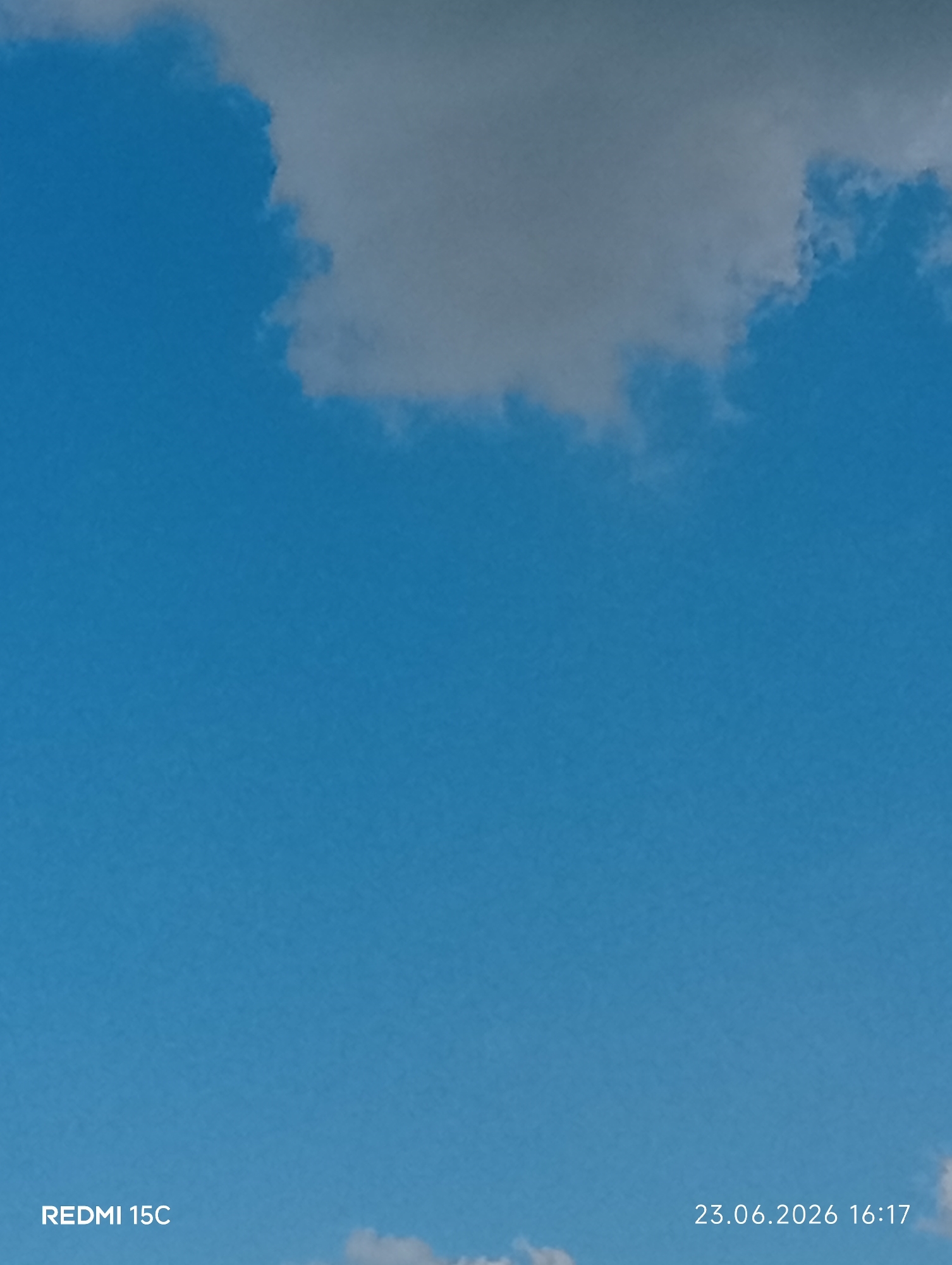The image shows a bright blue sky with scattered white clouds. The clouds are fluffy and appear to be cumulus clouds. The sky is clear and vast, indicating good weather. The lighting suggests it is daytime, likely mid-morning or afternoon, given the brightness and the soft shadows on the clouds. There are no people or man-made objects visible in the scene, focusing solely on the natural elements of the sky. The photograph appears to be taken from a ground-level perspective, looking upwards.
The bottom left corner of the image displays the text "REDMI 15C", which is likely a watermark indicating the device used to take the photo. In the bottom right corner, the date and time "23.06.2026 16:17" are visible, providing specific temporal information about when the image was captured. The combination of the date and the clear sky suggests a pleasant summer day. The location context of Mykolaivka, Ukraine, places the scene in Eastern Europe.