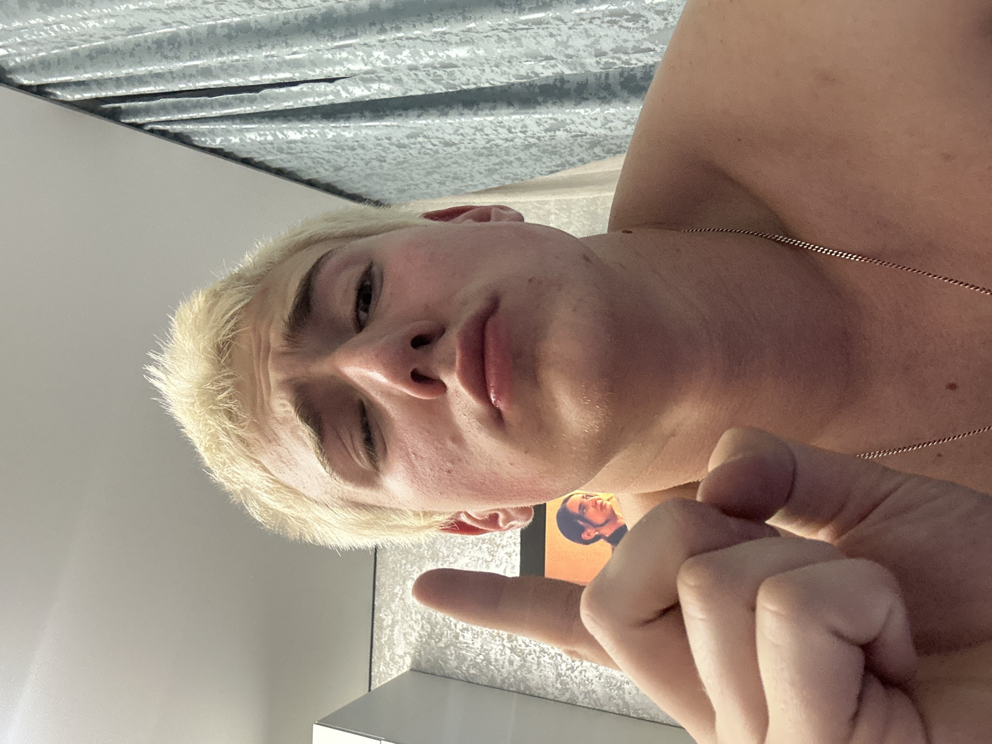 The image captures a close-up, rotated portrait of a person, likely a young adult male, with bleached blonde hair. The subject looks directly at the camera with a somewhat serious or contemplative expression, their lips slightly parted. The image is rotated 90 degrees clockwise, making the subject's head appear on the left side of the frame and their body extending to the right.
The person is shirtless or wearing minimal clothing, revealing a shoulder and chest. A thin, gold-colored chain necklace is visible around their neck. In the foreground, part of a hand is visible, with the index finger extended, possibly in a pointing or gesturing motion.
The setting appears to be indoors. In the background, to the subject's upper left, a textured surface resembling corrugated metal or patterned wallpaper is visible. To the lower right, a plain, light-colored wall forms part of the background. Behind the subject's arm, a small portion of what appears to be a poster or screen is visible, displaying a headshot of another individual with dark hair, rendered in warm, orange tones. There is no clearly legible text in the image.