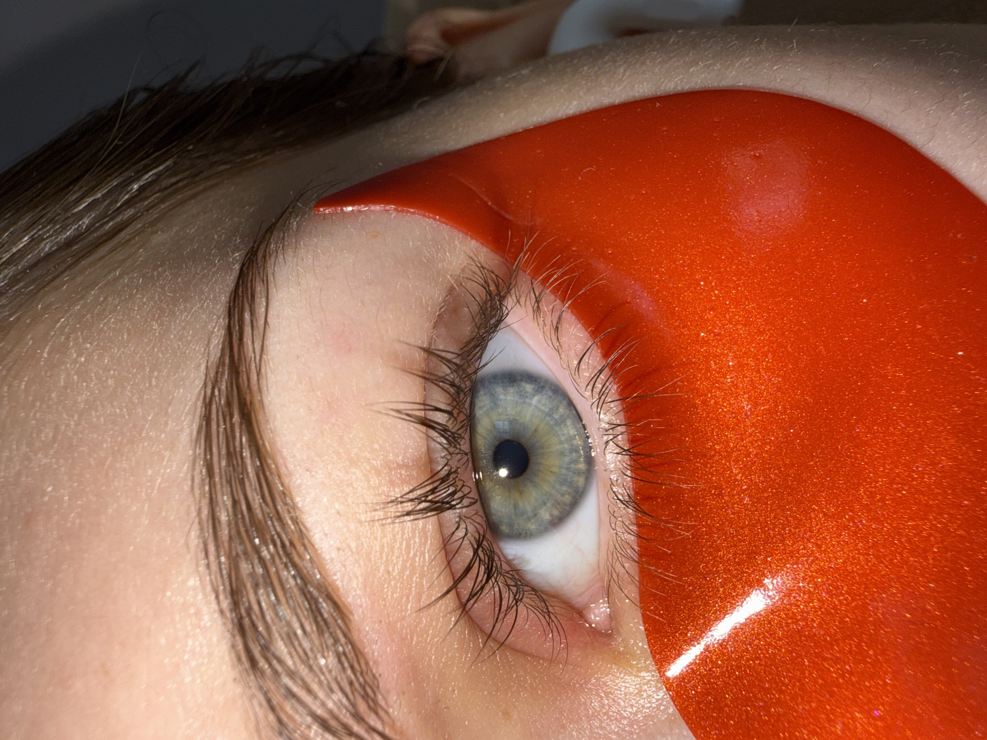This close-up image presents a person's left eye during an aesthetic beauty treatment, likely eyelash extensions or a lash lift, in Sofia, Bulgaria. The central focus is the eye, featuring a captivating light blue-green iris with a dark, round pupil. Long, dark upper eyelashes are prominent, while the lower eyelashes are gently secured and covered by a vibrant, glittery orange eye pad.
The orange eye pad is a curved, reflective accessory, possibly made of silicone or a gel-like material, designed to protect the lower lashes and skin during the procedure. It sits snugly along the lower eyelid, contrasting sharply with the fair skin tone. The skin around the eye appears smooth, with some fine hairs visible on the eyebrow and the area above.
A distinct reflection of a light source is visible on the surface of the iris and as a bright streak on the glossy orange pad, indicating bright artificial lighting, typical of a salon or treatment room. The person's gaze is directed upwards and slightly to the left, suggesting they are calmly engaged in the treatment. No text is visible in the image. The overall scene depicts a meticulous beauty procedure in progress.