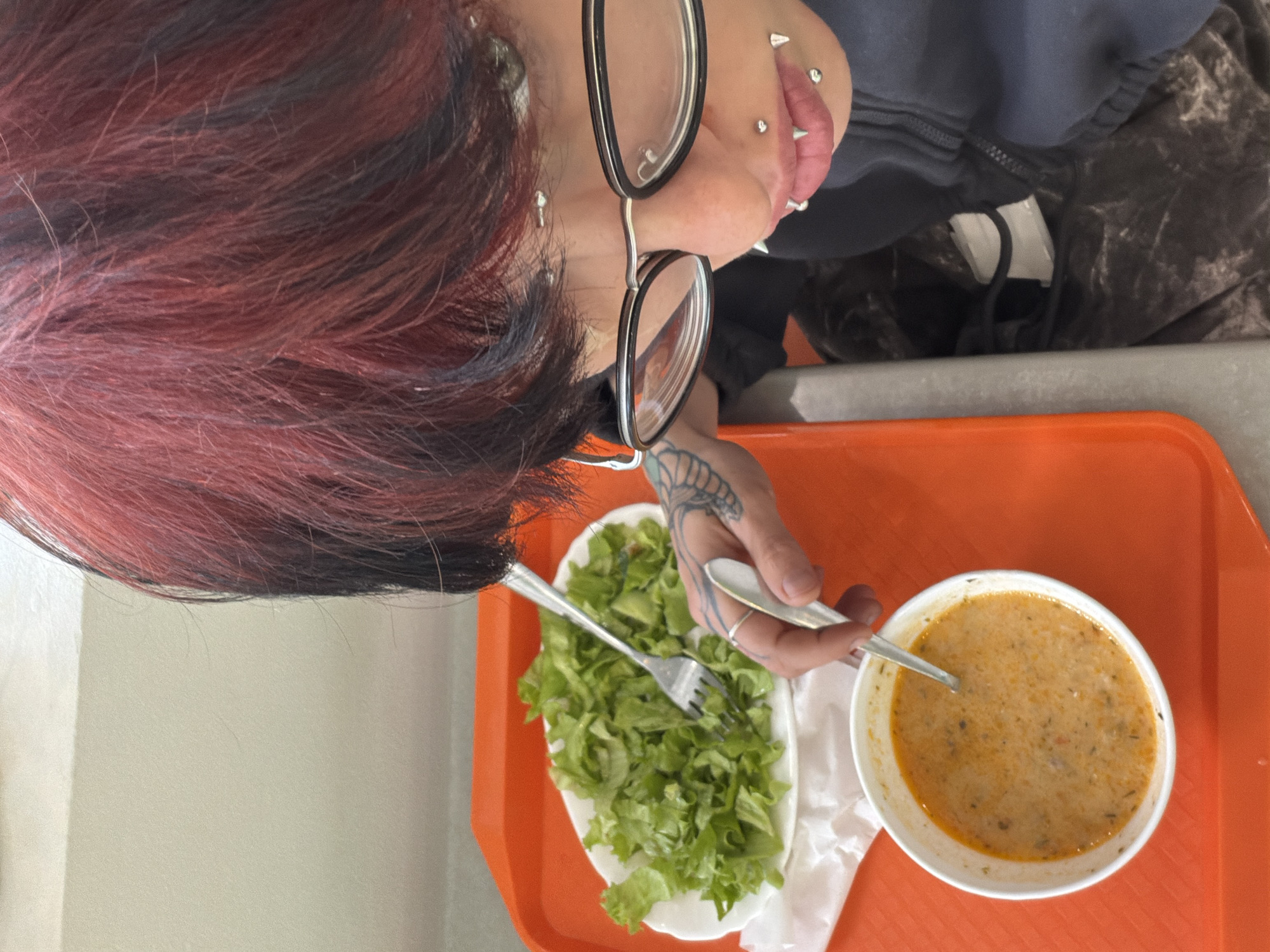 A person with red and black hair, wearing glasses, and sporting multiple lip piercings is positioned at a table. The individual is holding utensils in hand. An orange tray displays a white oval plate of lettuce salad and a white bowl filled with soup. The location is likely a food establishment, potentially in Sofia, Bulgaria.