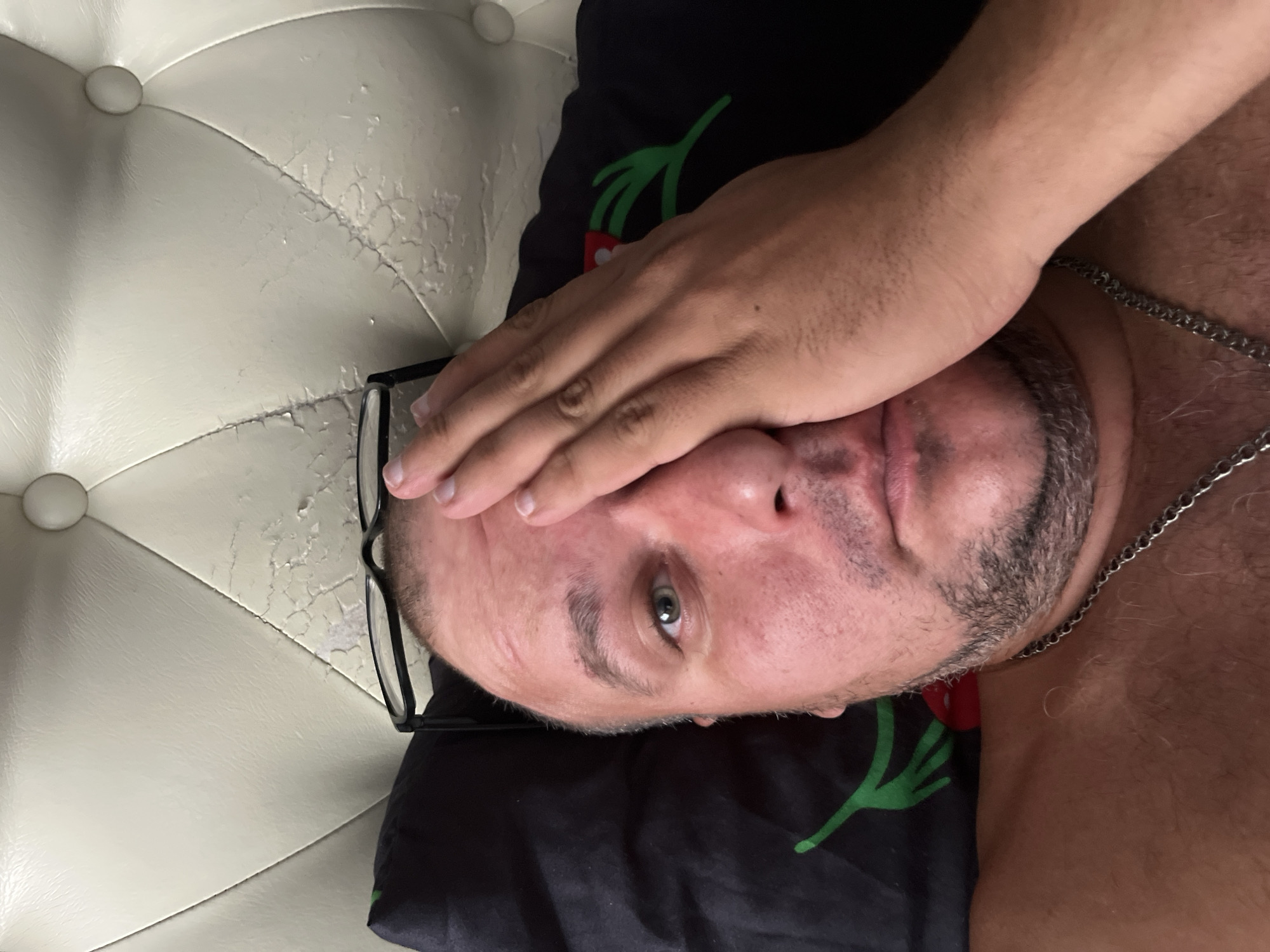 A man is lying in bed, covering his right eye with his hand. He is wearing a silver chain. The background shows a black pillow with a red and green design and a beige tufted headboard. The scene is indoors, likely a bedroom. No specific location in Minsk, Belarus is identifiable from the image.