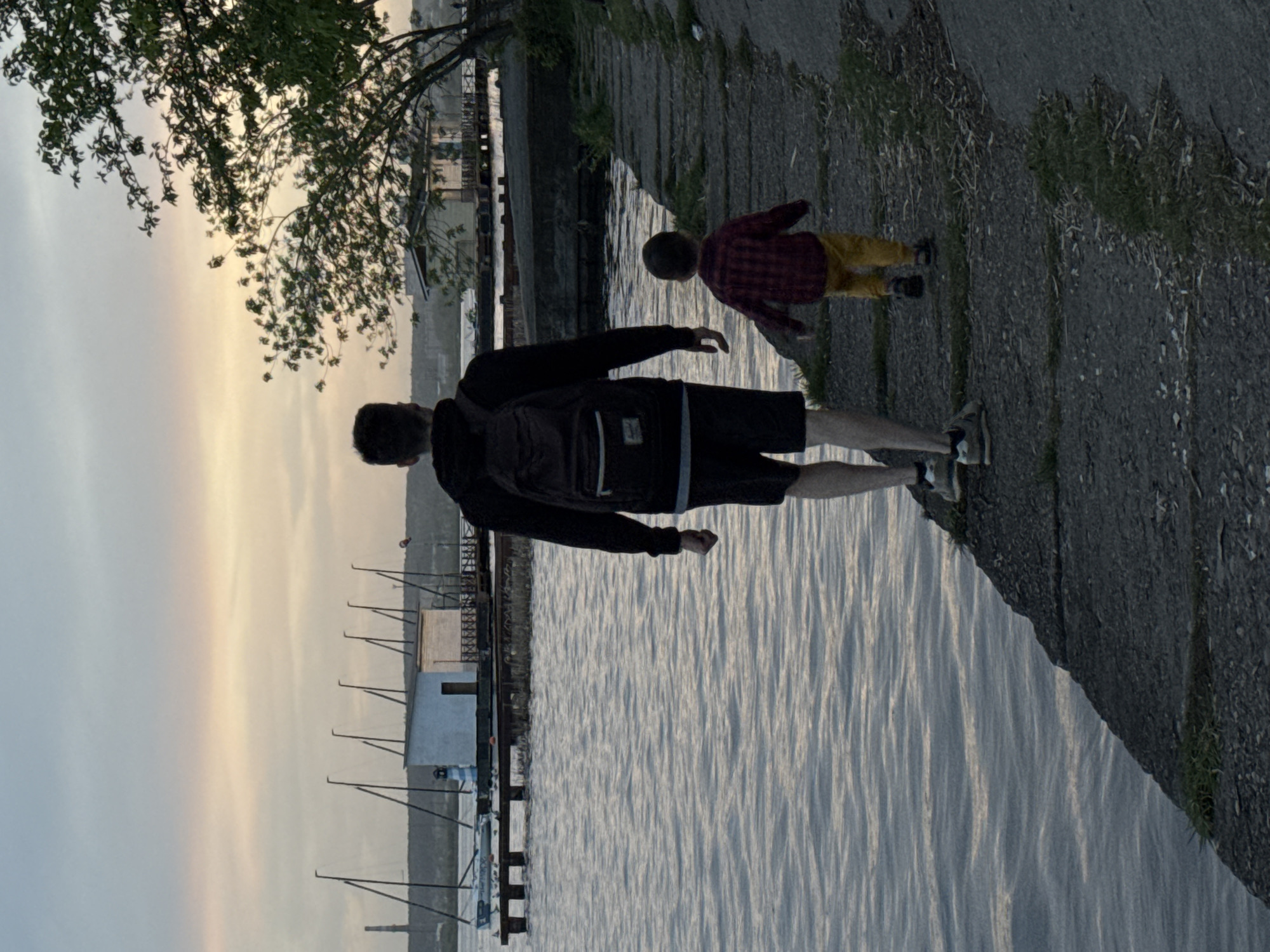 Two people, an adult male and a child, stand on a stone embankment next to a body of water. The man is wearing a dark jacket, shorts, and a backpack. The child is wearing a red and black plaid shirt and yellow pants. In the background, there are buildings, trees, and what appears to be a pier structure extending into the water. Given the presence of the pier and architectural style, the location is likely on the embankment of the Izhevsk pond in the city of Izhevsk, Russia.