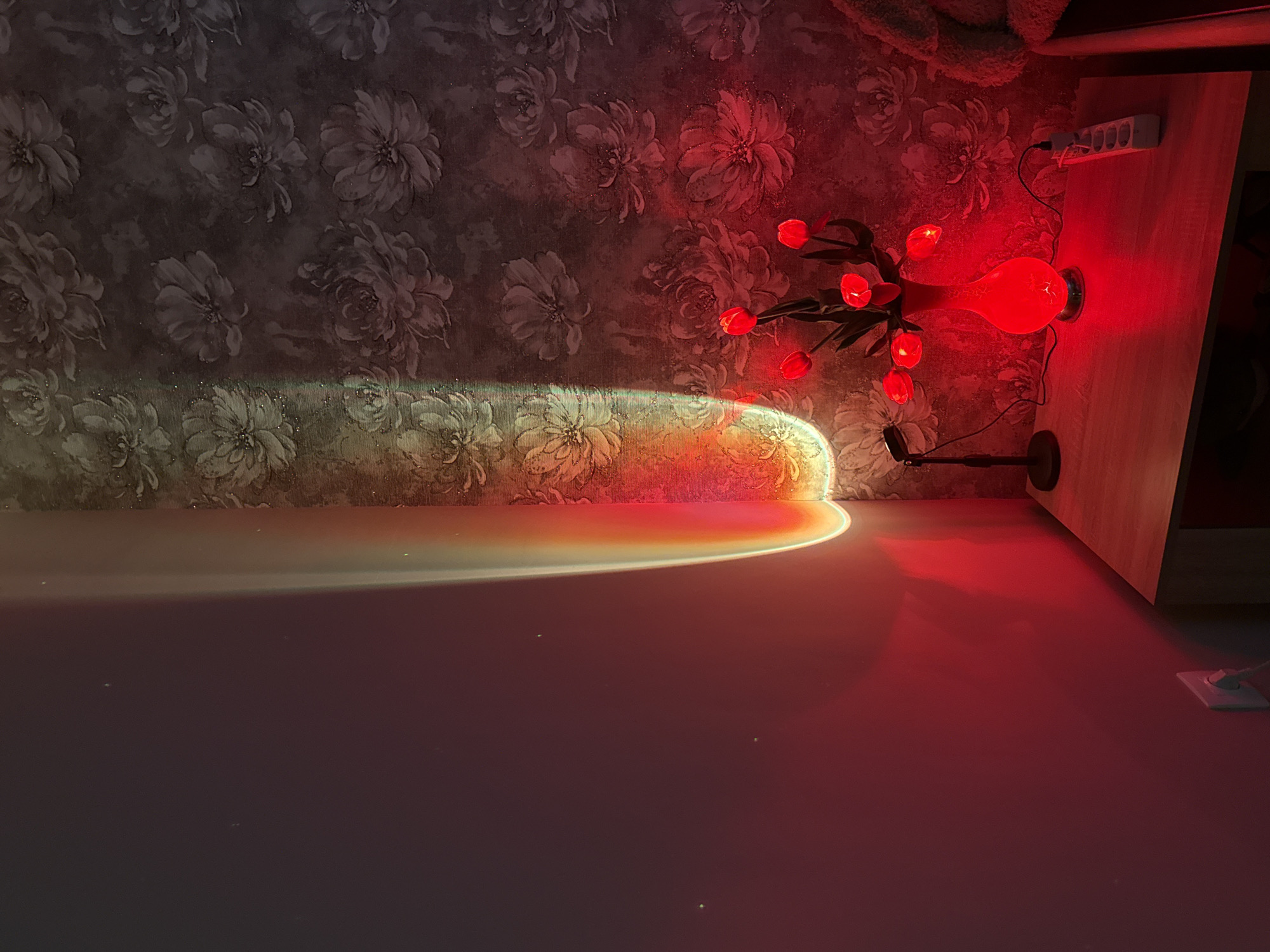 A sunset lamp casts a rainbow of light on a wall with floral wallpaper and a plain wall. A bouquet of red tulip-shaped lights in a red vase sits on a wooden nightstand. A power strip and a small black lamp are also on the nightstand. The room is dimly lit with a red glow.