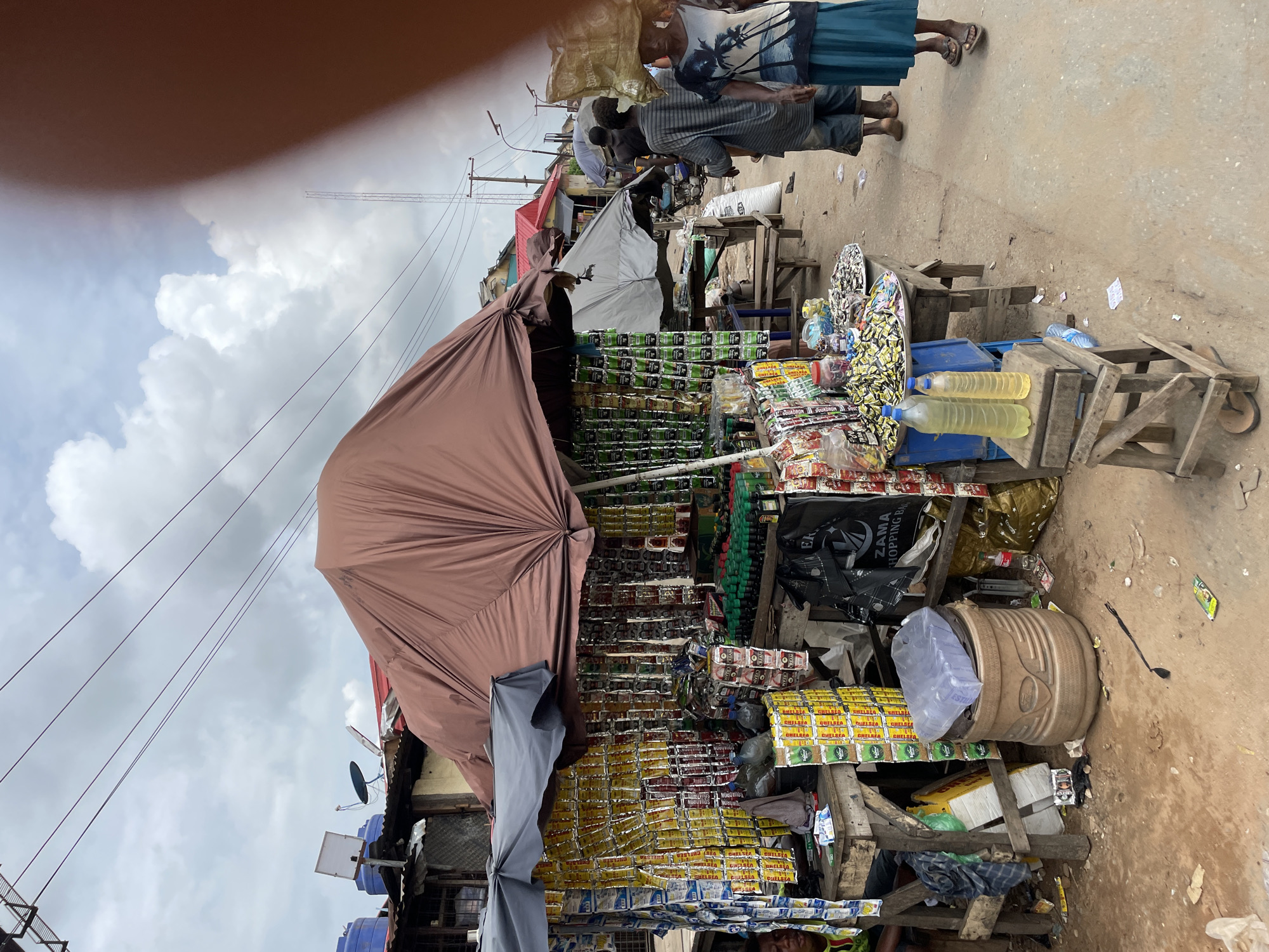The image depicts an outdoor market stall in what appears to be a city in an unknown location. The stall is laden with a variety of goods, including numerous small, individually packaged items hanging from the overhead structure and stacked on tables. These items appear to be food products or small consumer goods, with labels in various colors, predominantly yellow and green. Bottles of liquid, possibly cooking oil or fuel, are prominently displayed on a small wooden stool in front of the stall. To the right of the stall, several people are visible. One woman carries a large load on her head, while another person walks beside her. The ground is dirt and shows signs of wear and tear, with some scattered debris. The background reveals a street lined with buildings, some with corrugated metal roofs, and utility poles with overhead wires. The sky is overcast with clouds. The scene conveys a sense of a busy, functional marketplace.