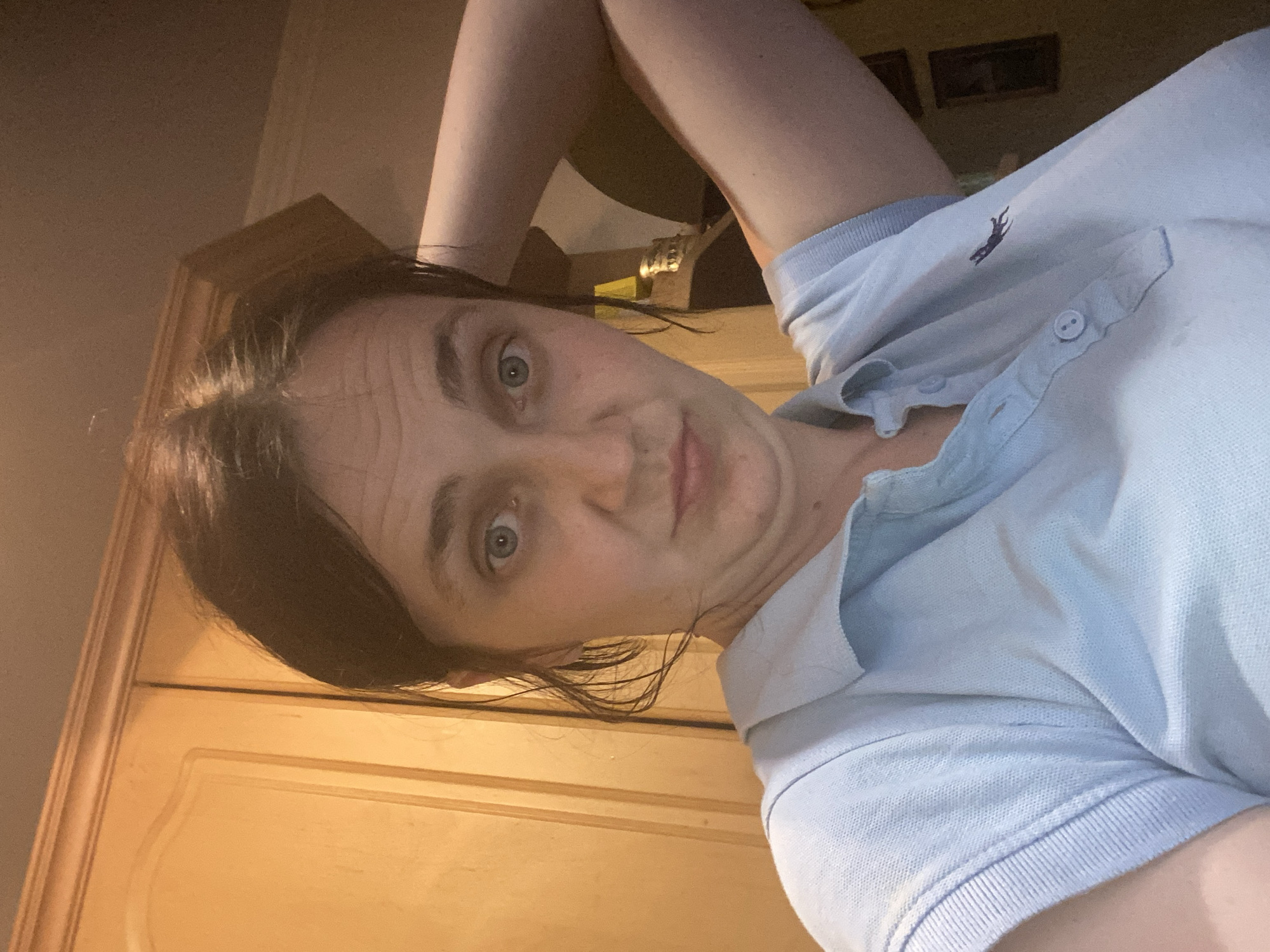 A woman, with dark hair, is looking at the camera. She has blue eyes and is wearing a light blue polo shirt with a small dark logo of an animal on it. Her right arm is bent and her hand is resting on the back of her head. The background appears to be a kitchen with wooden cabinets. The lighting is warm and appears to be artificial. There are two framed pictures on the wall in the background.