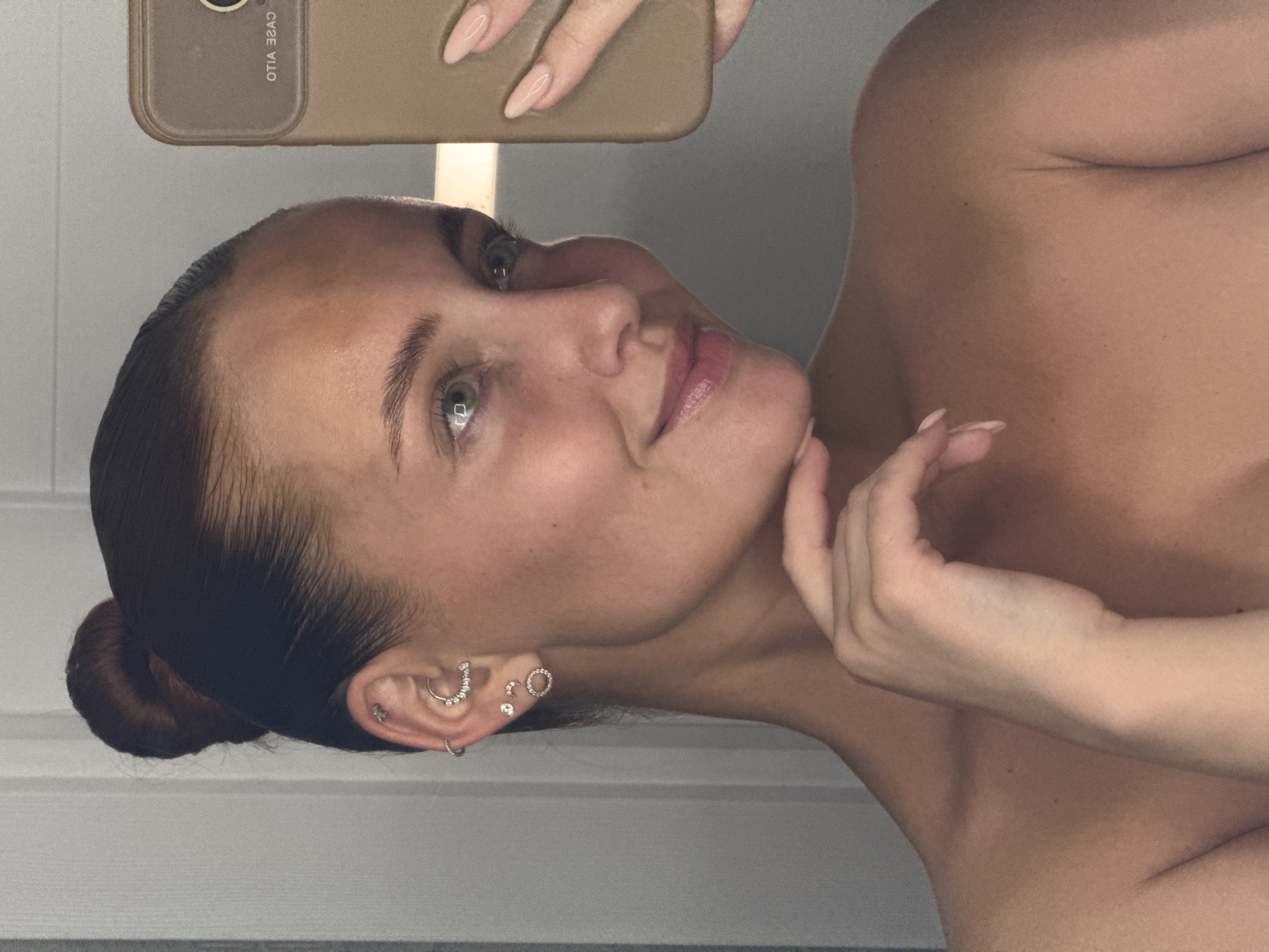 The image captures a close-up selfie of a woman, oriented sideways, with her head tilted to the right. She looks directly towards the camera with a soft, gentle smile, her light-colored eyes appearing green or hazel. Her dark hair is neatly pulled back into a bun. Her left hand, with light-colored manicured nails, holds a smartphone with a brown case above her head. The phone's screen reflects the reversed text "OTIA ƎSAC," which correctly reads "CASE VILO." Her right hand gently rests on her chin.
The woman appears to be bare-shouldered, with a portion of her chest visible. Her left ear is adorned with several small, intricate piercings, including a small hoop, a larger hoop studded with stones, and multiple tiny studs. The background is a plain, light-colored wall or surface, indicating an indoor setting. The lighting is soft and even, highlighting her features. The overall impression is one of calm and subtle introspection.