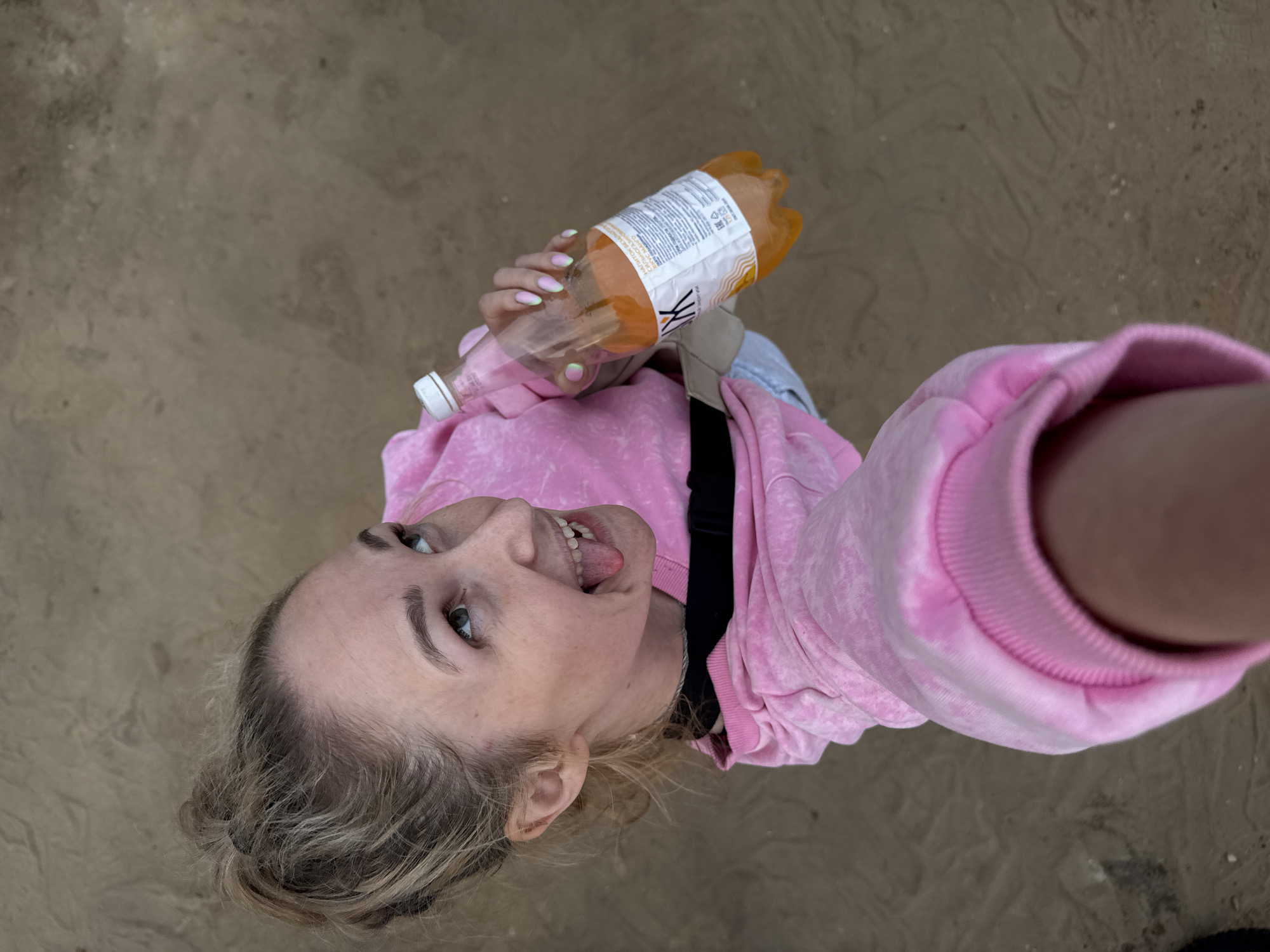 A woman is taking a selfie with a bottle of orange soda in Chaikovsky, Russia. She is wearing a pink hoodie and has her tongue sticking out. The background is sand.