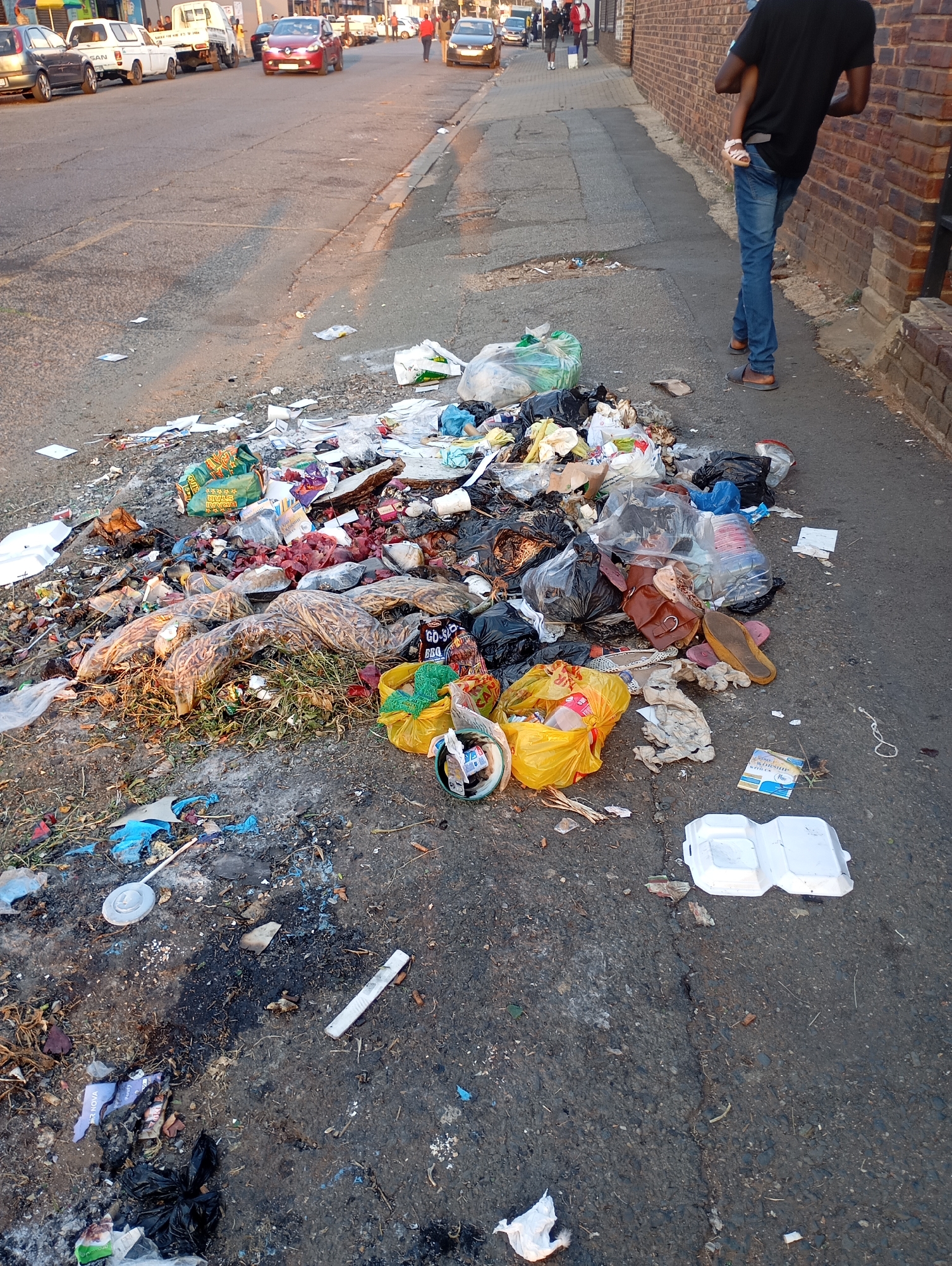 The image depicts a large pile of trash on a sidewalk in Johannesburg, South Africa. The trash includes plastic bags, food wrappers, discarded clothing, and general refuse. The sidewalk is cracked and stained. A man walks along the sidewalk, stepping around the trash. In the background, cars are parked along the street, and other pedestrians are visible. The environment suggests an urban, possibly low-income, area.