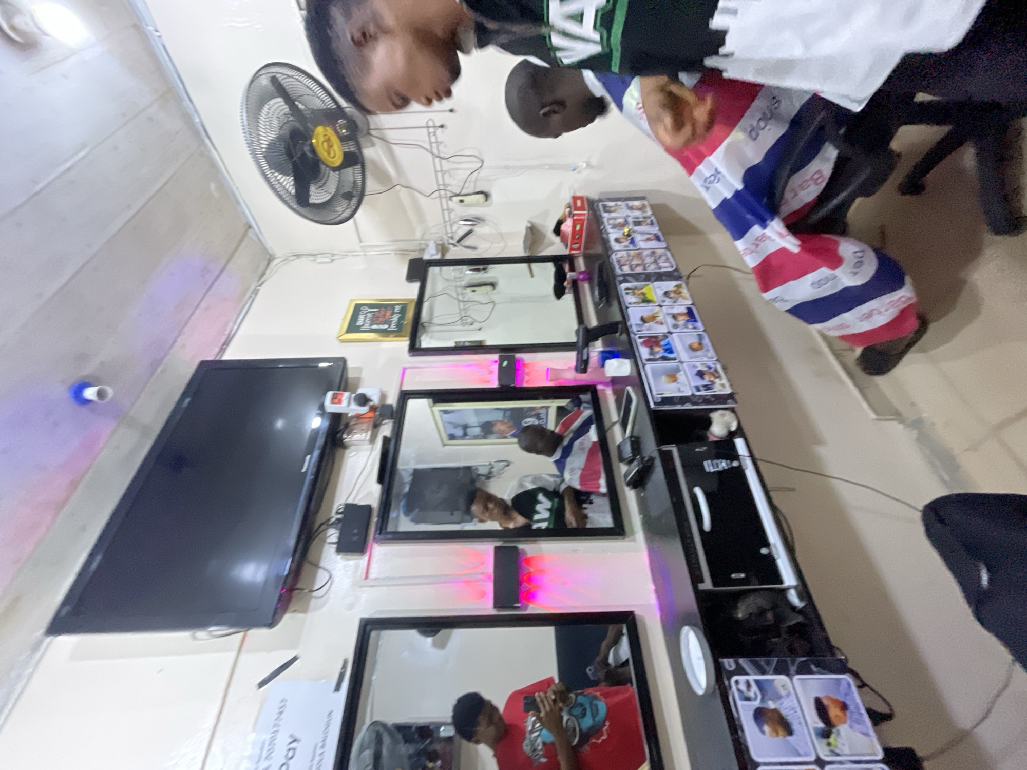 Inside a barber shop, presumably in Ado, Nigeria, a barber attends to a seated customer draped in a red, white, and blue barber cape. The shop features a flat-screen TV mounted on the wall, a fan, a row of mirrors, and a black shelving unit displaying various barbering tools and supplies. A collection of cards or pictures is lined up on the shelves. The setting suggests a small, functional space tailored for providing barber services.