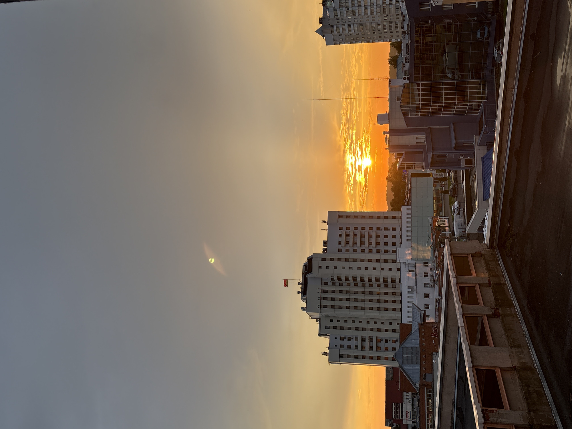 A medium-shot, vertical, daytime photograph captures a city skyline in Vitsebsk, Belarus, under a warm, hazy sky, indicating either sunrise or sunset. Dominating the scene is a tall, white, multi-story building with numerous square windows, adorned with a red and white flag at its peak. To the right, another high-rise building, light gray and segmented, rises towards the sky. In the foreground, a rooftop with a weathered, dark surface and a concrete parapet with small window openings suggests a vantage point for observation. Below, various commercial and residential buildings, including a modern structure with a glass facade, form the urban landscape. The sky is a gradient of soft grays at the top, transitioning to a vibrant orange glow near the horizon, where the sun is partially obscured by textured clouds, casting a warm light across the city. The presence of streetlights, antennas, and parked vehicles on the streets below further define the urban setting. No individuals are clearly visible in the frame, but distant silhouettes on the roof of the white building might suggest human presence.