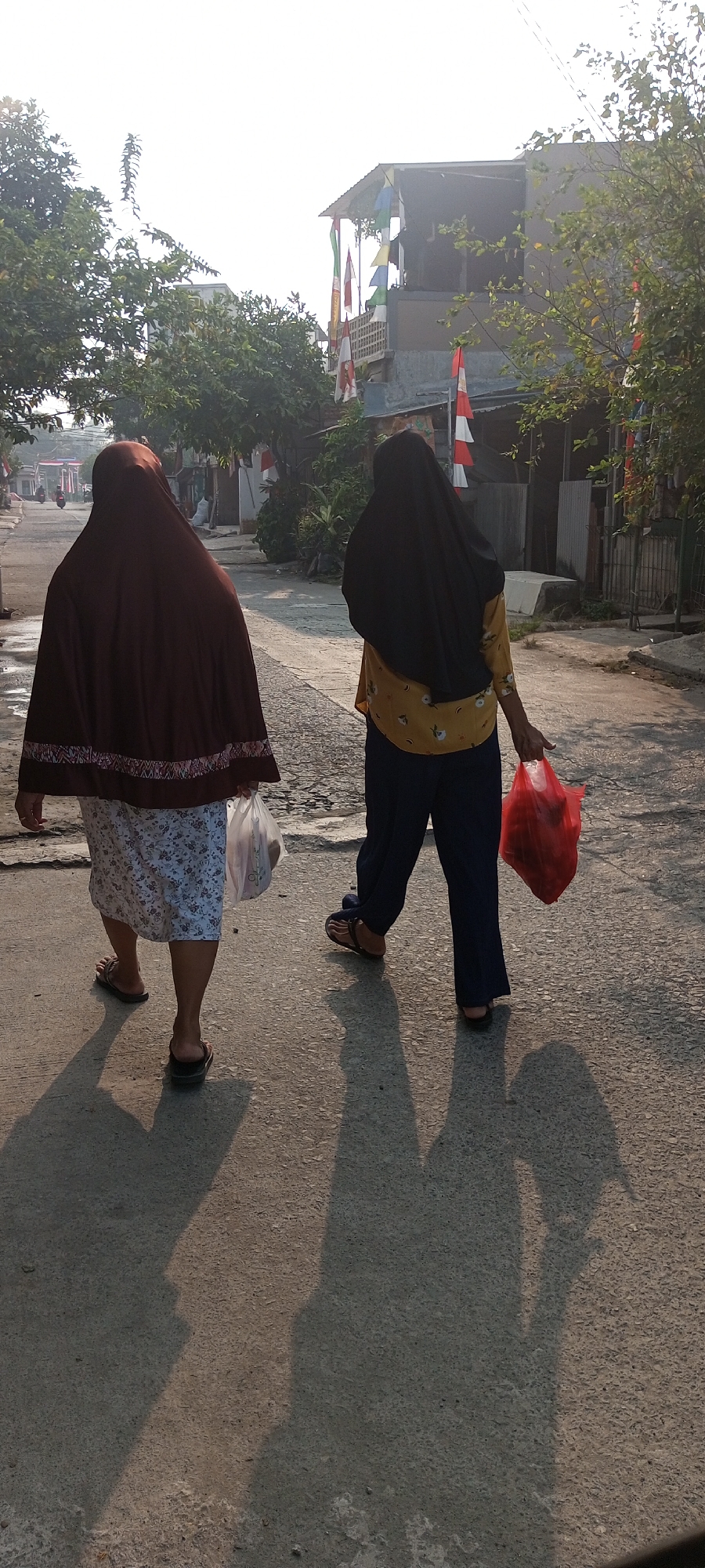 In Setu, Indonesia, two women are walking away from the camera on a paved road. The woman on the left wears a dark brown headscarf and a patterned skirt, carrying a white plastic bag in her right hand. The woman on the right has a black headscarf and wears a yellow patterned shirt with dark blue pants, carrying a red plastic bag in her right hand. Both are wearing sandals. The road is lined with houses and trees, and Indonesian flags are visible on some buildings, suggesting a national holiday or celebration. The sun is casting long shadows on the road, indicating it is either early morning or late afternoon.