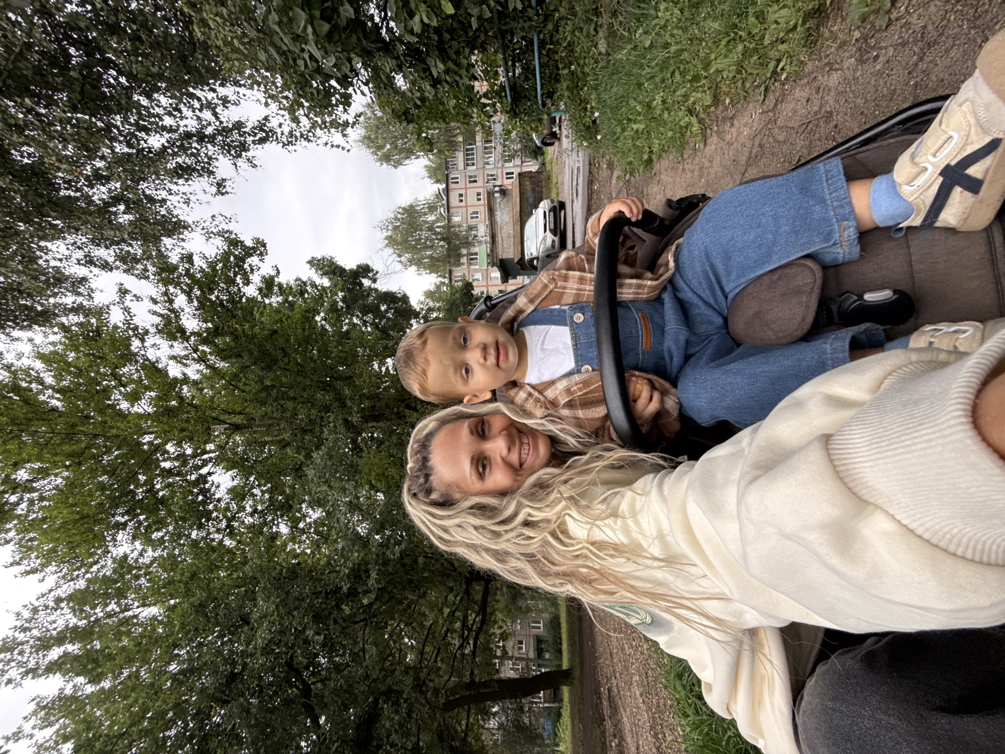 In Rusanovo, Russia, a woman and a young child are in a stroller on a path. The woman, with blonde hair pulled back, smiles at the camera while the child, wearing a plaid shirt and denim overalls, looks forward. The background shows apartment buildings and trees.
