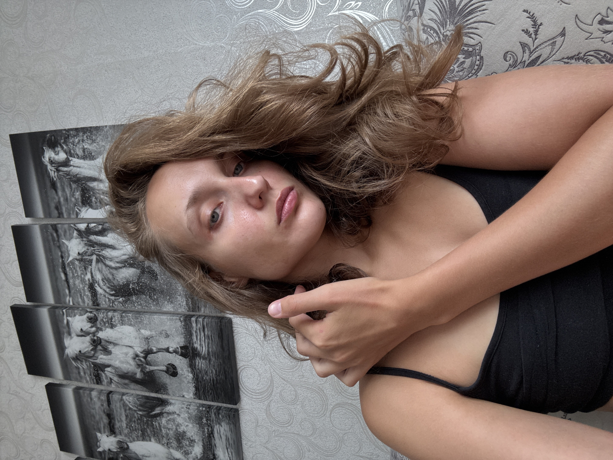 A young woman with long, wavy blonde hair is positioned in a close-up shot, looking directly at the camera. She has light skin with a touch of blush on her cheeks and light-colored eyes. She is wearing a black tank top. Her left arm is raised and her hand is gently touching her neck. In the background, there are four framed black and white pictures of horses running through water. The wall behind the pictures has a subtle, light-colored patterned wallpaper. The overall setting appears to be indoors, possibly a living room. The image does not contain any information that would allow for a specific location within the city of Kuyeda, Russia to be identified.
