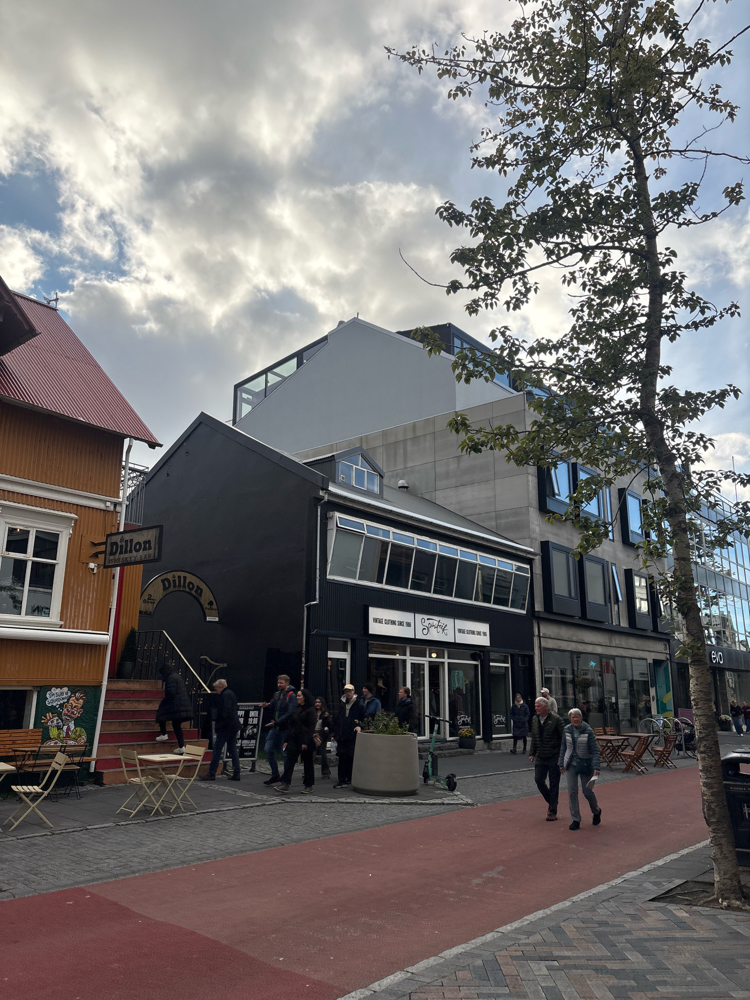 The image shows a street scene in Reykjavik, Iceland, likely on Laugavegur street, a popular shopping and entertainment area. In the foreground, a red-painted pedestrian walkway runs alongside a brick-patterned sidewalk. Several outdoor tables and chairs are set up on the sidewalk.
On the left side of the image, an orange building with a red corrugated metal roof features a window with a sign that reads "Dillon Whiskey Bar." Further down the street, a black building with a curved entrance and a sign that says "Dillon" is visible. A set of red carpeted stairs leads up to the entrance. Several people are seen walking on the sidewalk, some entering or exiting the black building.
To the right of the black building, a modern gray building with multiple floors and large windows dominates the mid-ground. Below this, a storefront with large glass windows has signs for "Vintage Clothing Since 1995" and "Spútnik." Further to the right, another building has the word "eva" visible on its facade.
A tall tree with green leaves stands on the right side of the street, partially obscuring the view of the buildings behind it. The sky is partly cloudy with patches of blue visible. The overall atmosphere suggests a bustling urban environment.