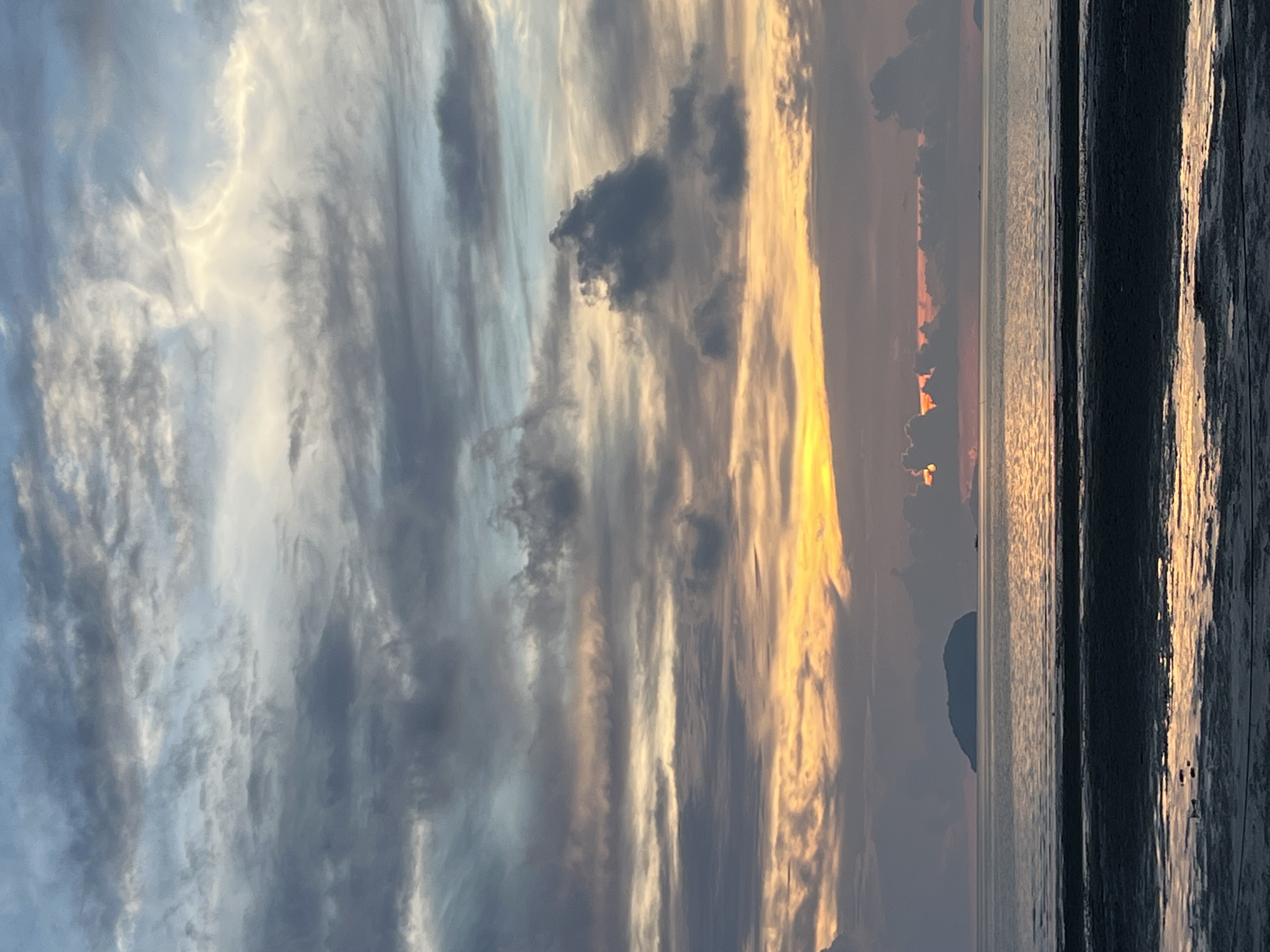 The image depicts a serene sunset over the ocean, likely taken from a beach in Cuyo, Philippines. The sky is a canvas of dramatic clouds, transitioning from pale blues and greys at the top to warm oranges and yellows closer to the horizon. The setting sun is partially obscured by dark clouds, casting a golden glow on the water's surface. In the distance, a small, uninhabited island stands silhouetted against the vibrant sky. The foreground shows a wet, sandy shore, reflecting the colors of the sunset. No people or specific structures are visible, emphasizing the natural beauty of the scene.