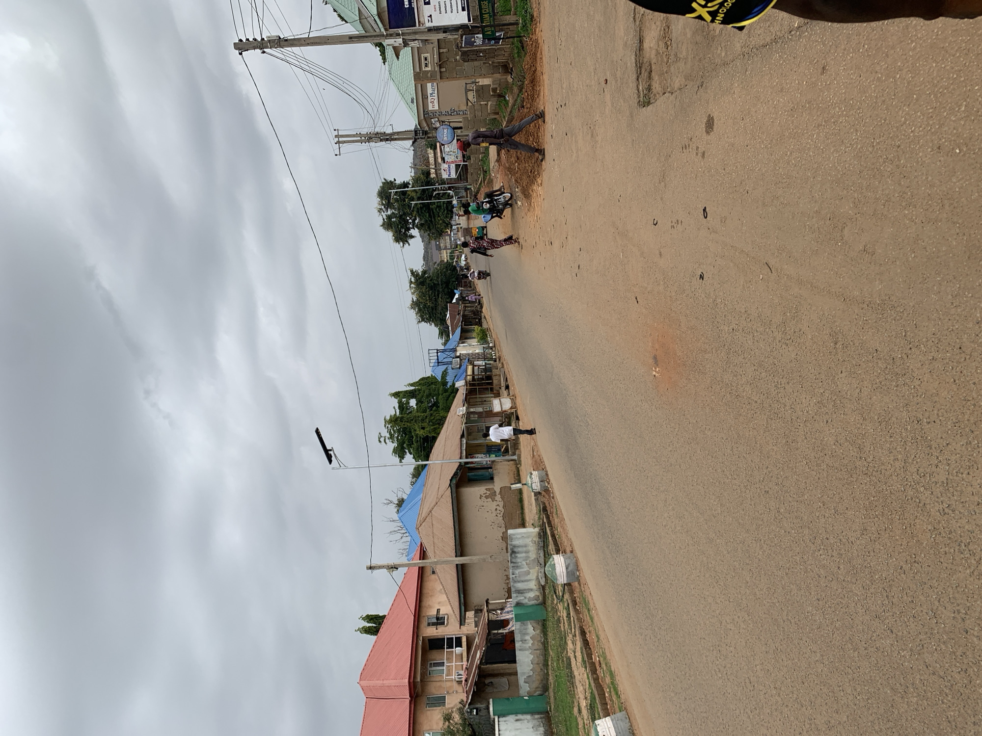 The scene depicts a street in New Stadium, Nigeria, under an overcast sky. The road, made of brown asphalt, stretches into the distance, slightly curving to the left. Several individuals are walking on or near the road, some appearing to cross it. A few motorcycles are visible further down the street. Buildings of varying sizes and colors line the left side of the road. Utility poles with wires run along the street. A streetlight is also present on the left side. The foreground shows a glimpse of a person wearing a yellow and black object, partially cropped out of the frame. The environment appears to be a mixed residential and commercial area.