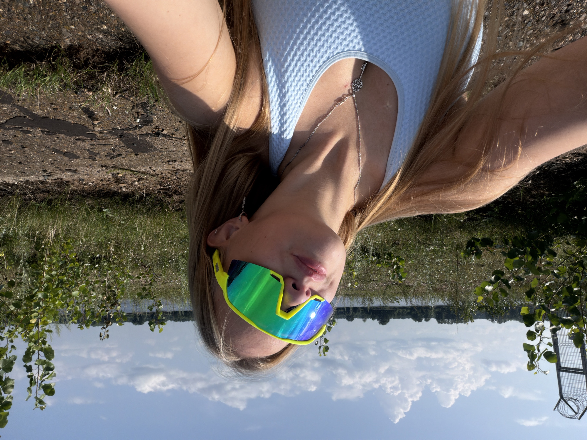 A young blonde woman is taking a selfie outdoors with a body of water and trees in the background. She is wearing bright yellow-green cycling glasses, a light blue tank top, and a silver necklace with a key pendant. The ground is a mix of dirt, gravel, and patchy grass. A metal fence with barbed wire is visible to the left of the frame. The sky is blue with scattered clouds. The scene appears to be near the Kama River in the city of Chaikovsky, Russia.