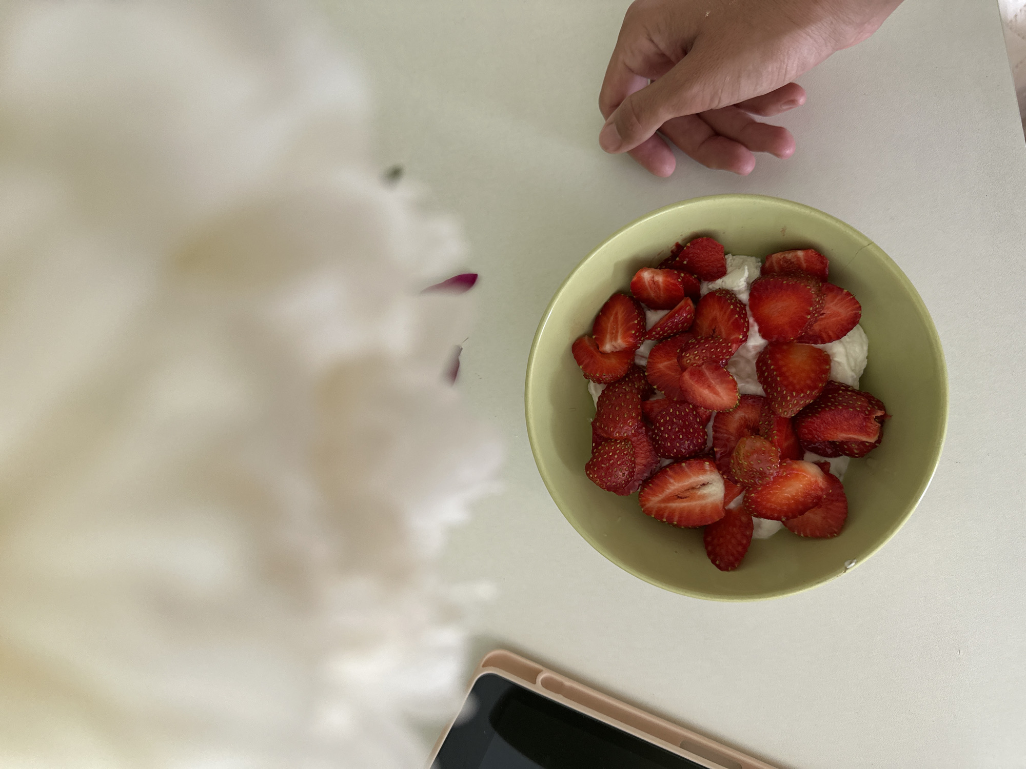 A top-down, close-up shot shows a light green bowl filled with sliced strawberries and a white creamy substance, likely yogurt or whipped cream. The bowl is placed on a white surface, possibly a table. To the left of the bowl, a smartphone in a light pink case is visible. To the right of the bowl, a person's hand with extended fingers is reaching towards the food. In the background, out of focus, is a large, soft, white object, possibly a flower or fabric. The overall scene is bright and appears to be indoors. There is no visible indication of the location in Chaikovsky, Russia.