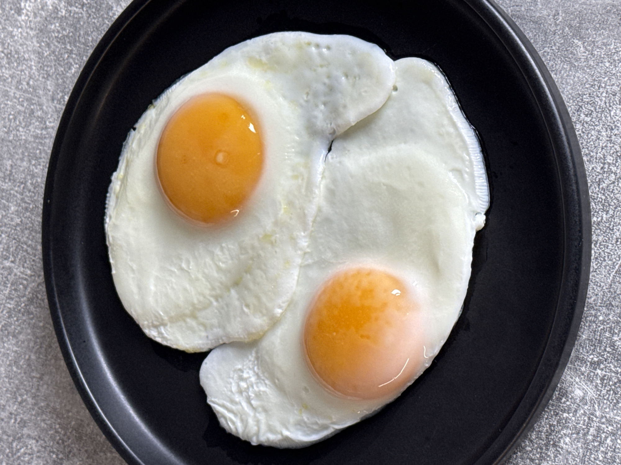 An overhead, close-up shot features two sunny-side-up fried eggs presented on a black, round plate. The plate, which has a matte or slightly textured finish and a subtle rim, rests on a grey, speckled surface that appears to be a countertop or table.
Both eggs are cooked with fully set, white egg whites and vibrant orange yolks. The yolks appear slightly glossy and plump, suggesting they are still soft or runny. The egg whites show some slight bubbling and crisping around the edges, a common characteristic of frying. The two eggs overlap slightly in the center of the plate, with one positioned towards the top left and the other towards the bottom right. The lighting is even, highlighting the textures of the egg whites and the distinct colors of the yolks. No people or visible text are present in the image. The scene suggests a prepared meal, possibly breakfast or brunch, ready for consumption.