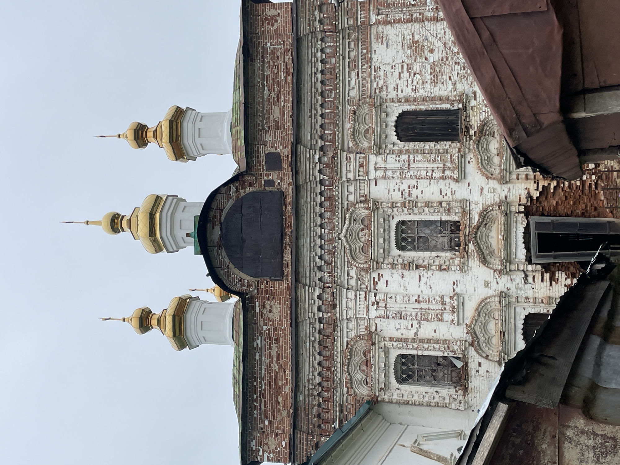The image shows the exterior of an old, weathered, white-washed brick church with a red tiled roof and golden domes. The church features multiple arched windows, some with bars and broken glass, and decorative brickwork. The facade is partially covered by a dark, weathered metal sheet. In the foreground, there are two dark, sloped roof structures made of corrugated metal, partially obscuring the lower part of the church. The sky is overcast and gray. The scene appears to be in the village of Кстинино, Russia, indicated by the context provided. There are no people visible in the image.